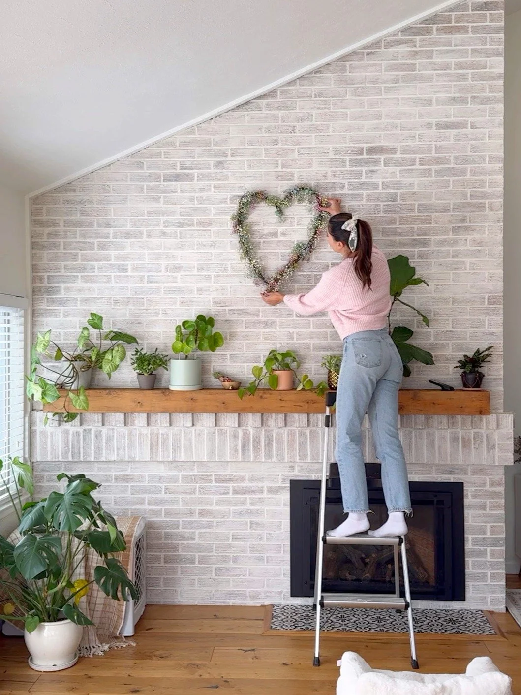 Woman standing on a step stool hanging a heart-shaped floral wreath on a white brick wall with potted plants on a wooden mantle and a large monstera plant in a white pot nearby.