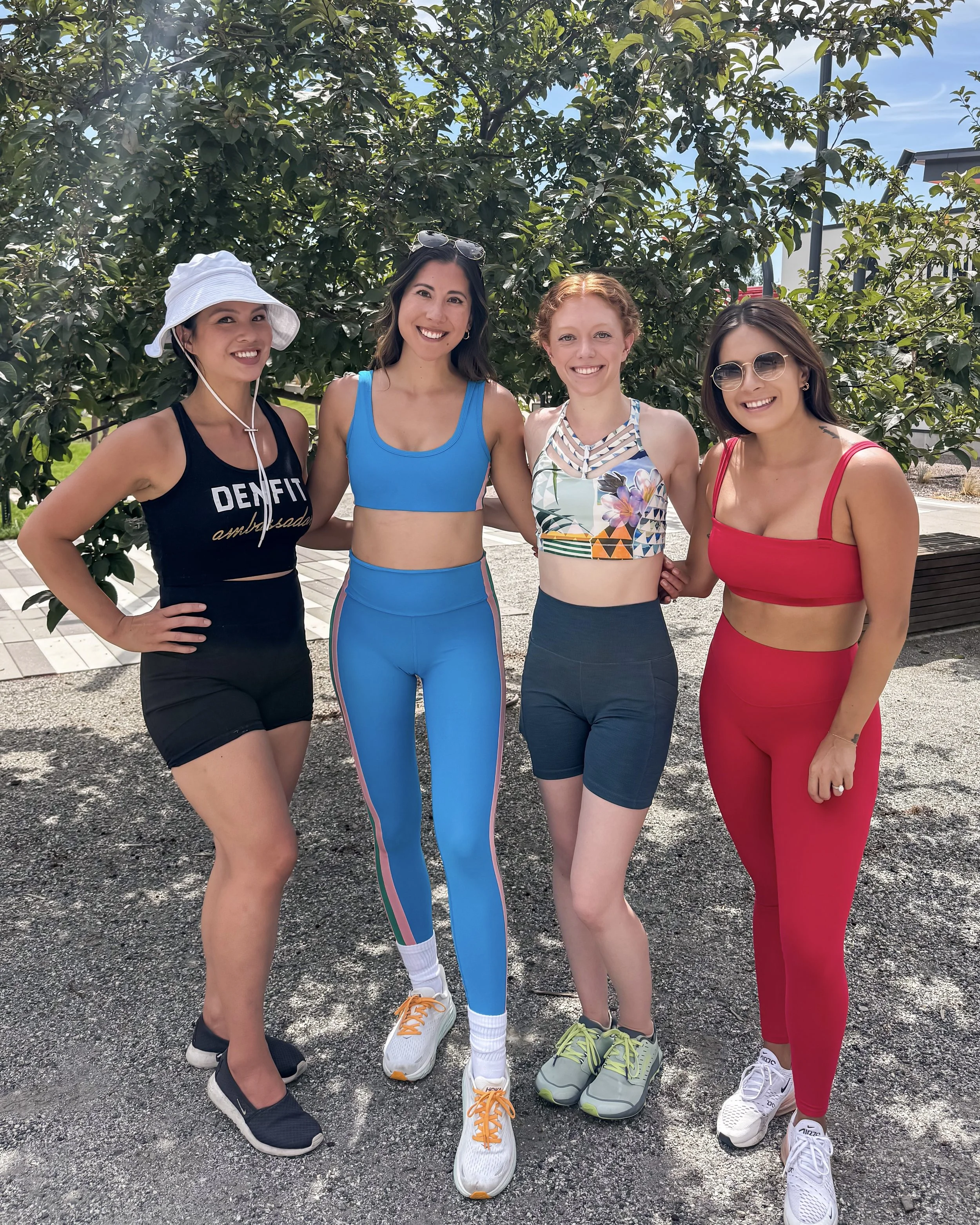 Four women standing outdoors in a line, smiling, with green trees behind them. They are dressed in colorful athletic clothing and sneakers.