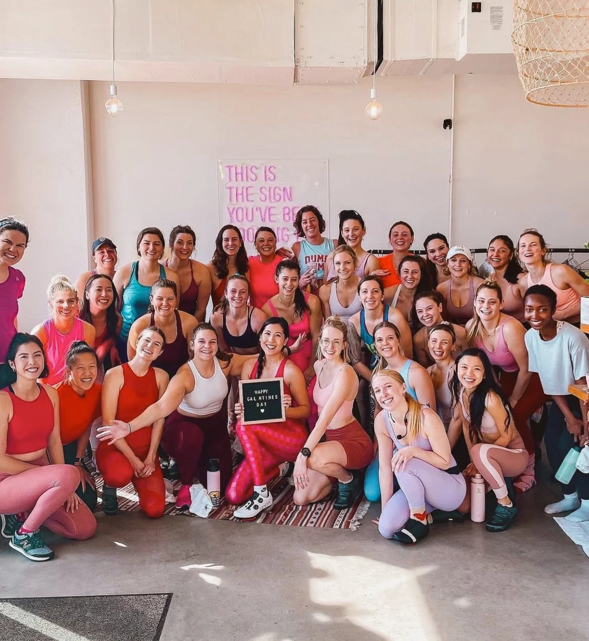 Group of women in workout clothes smiling together in a fitness class or gym, with a sign that reads 'Happy Galentine's Day.' Some women are kneeling or sitting in front, and others are standing behind.
