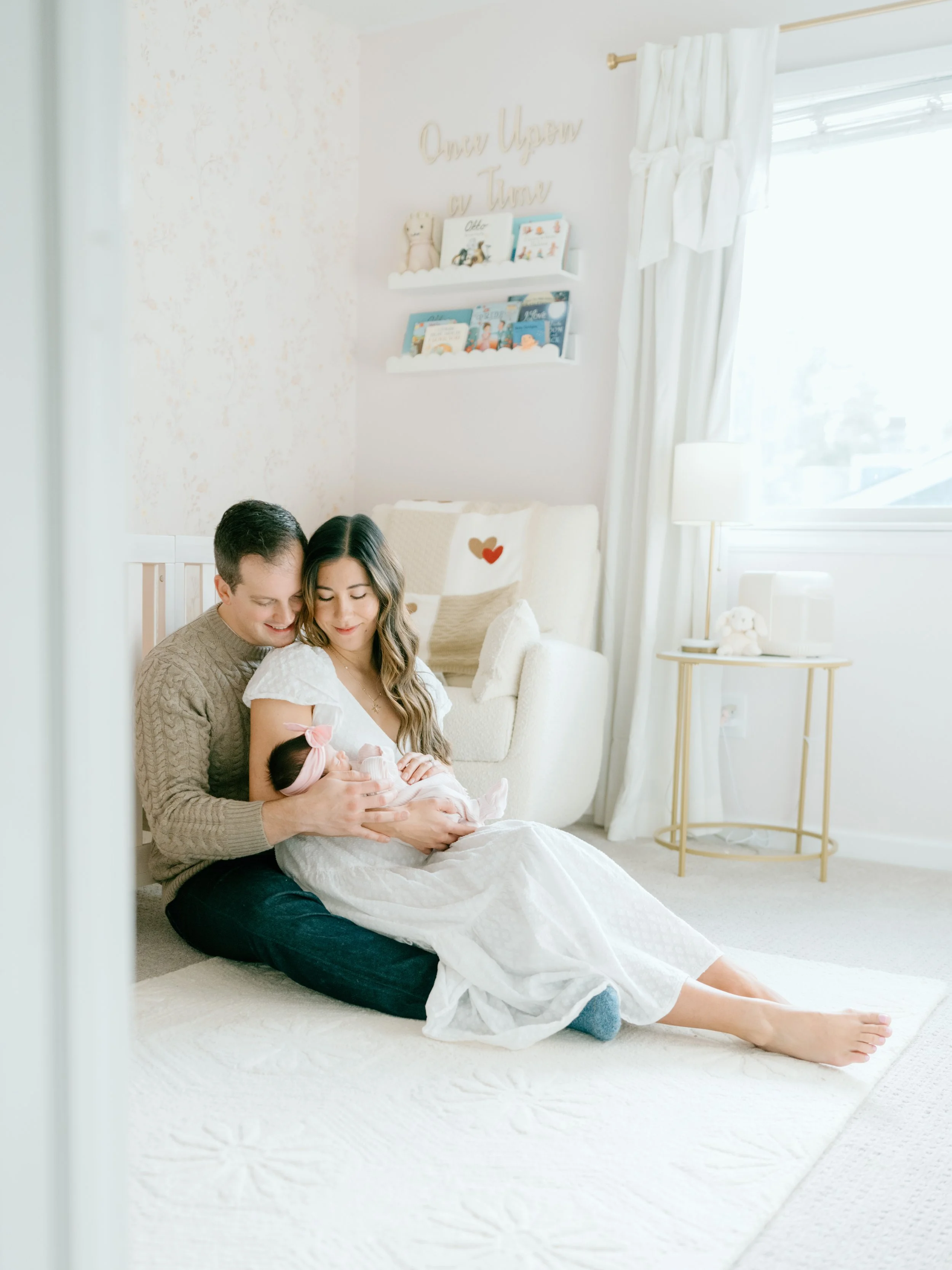 A family sitting on a white rug in a bright nursery, holding a newborn baby girl with a pink bow, with a beige armchair behind them and cozy decor on the wall.