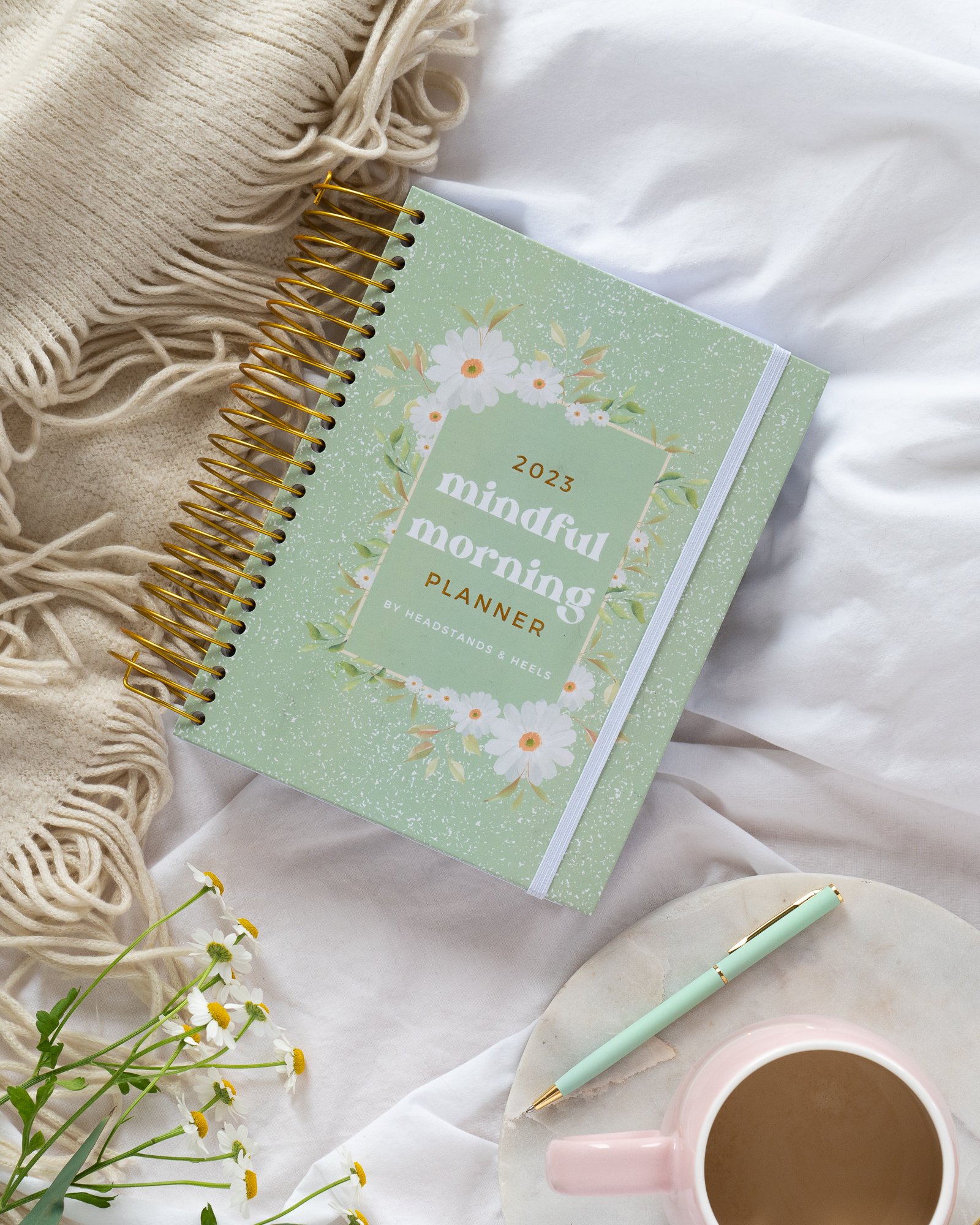 2023 mindful morning planner with floral cover, a pen, a cup of coffee, and daisies on a white bed with a beige blanket.