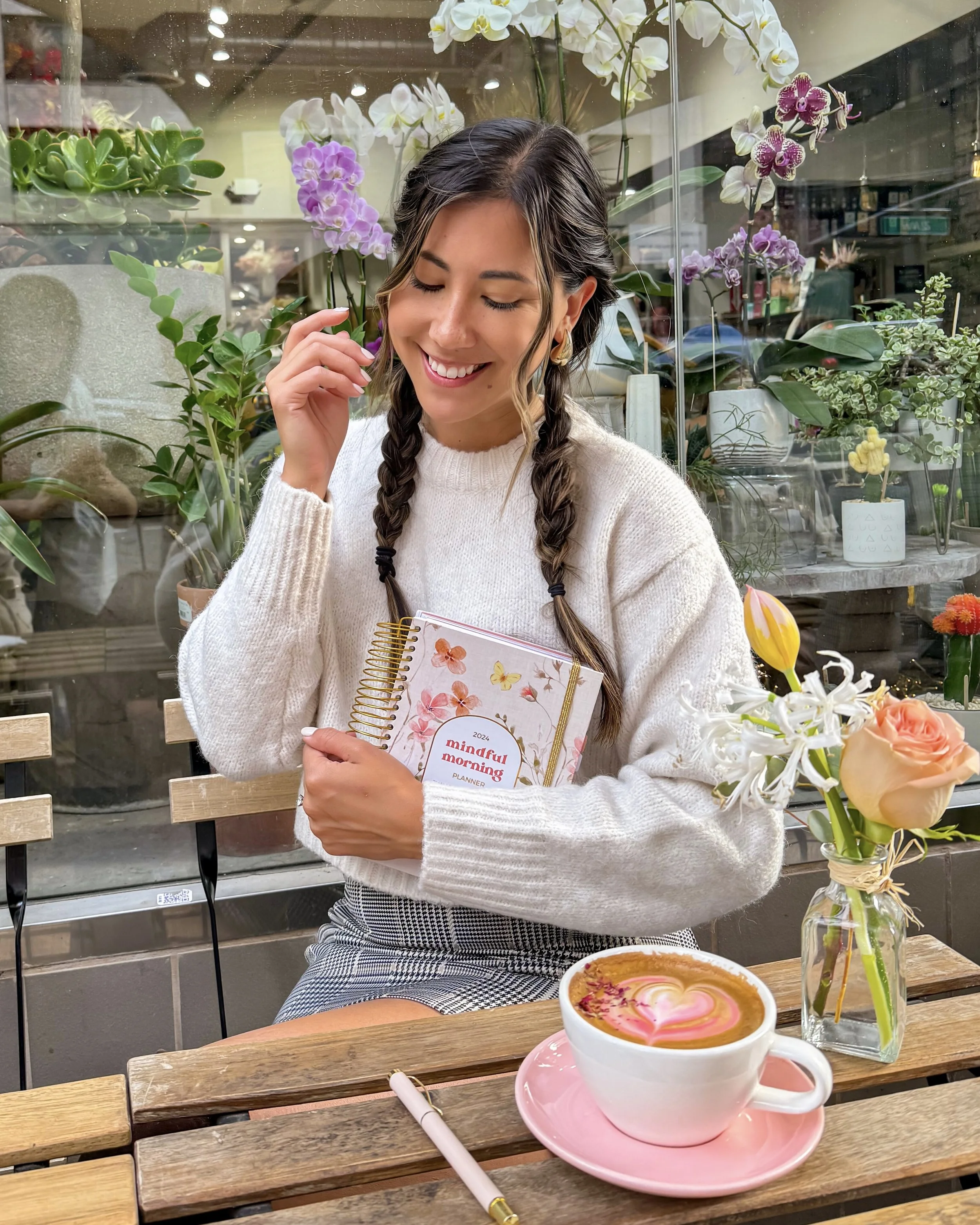 A smiling woman with braids holding a planner, seated at a wooden table in a flower shop or cafe, with a latte decorated with a heart in a white cup on a pink saucer, a pen, and a vase of flowers on the table, surrounded by various potted plants and orchids.