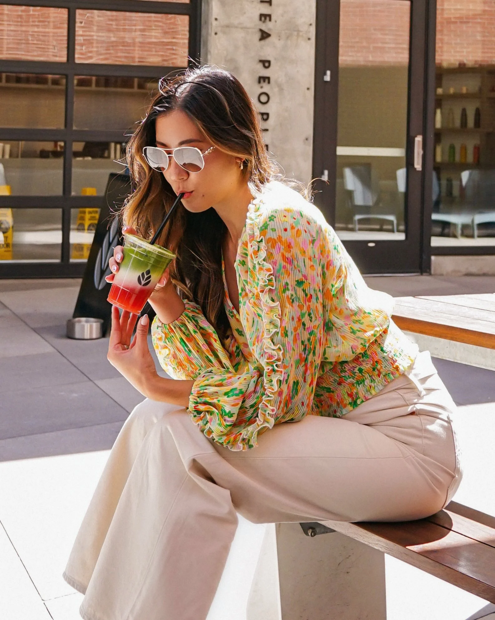 Woman sitting outdoors on a bench, drinking a layered green and red beverage through a straw, wearing sunglasses, a colorful blouse, and beige pants.