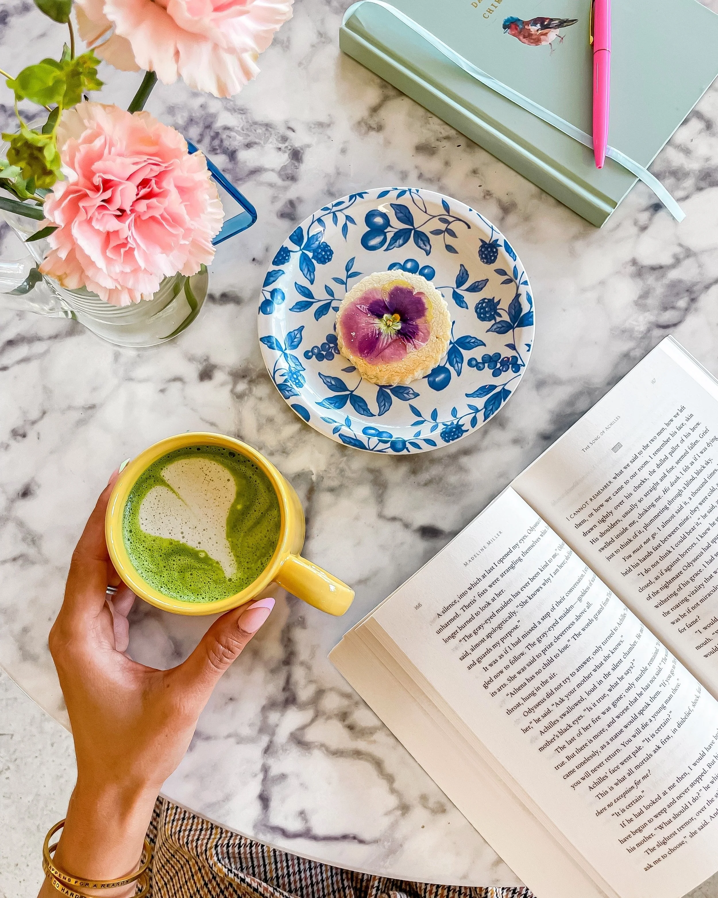 A marble table with a pink carnation flower in a glass jar on the left, a hand holding a yellow mug with green tea or matcha, an open book on the bottom right, a decorative plate with a cupcake topped with edible flower petals in the center, a book with a pink pen and a green notebook with a bird on the cover on the upper right.