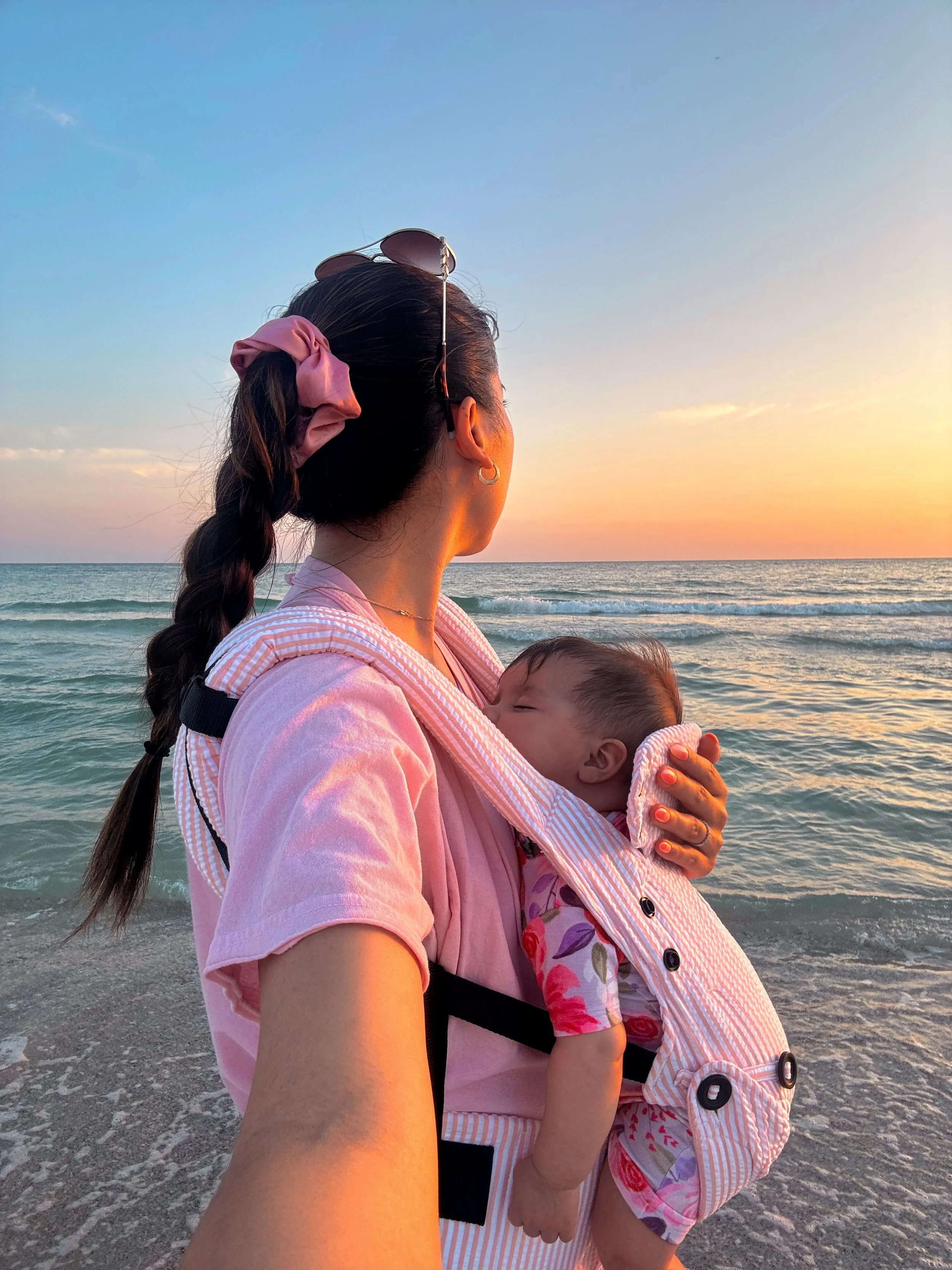 Woman in pink shirt carrying a sleeping baby in a striped carrier on a beach during sunset.