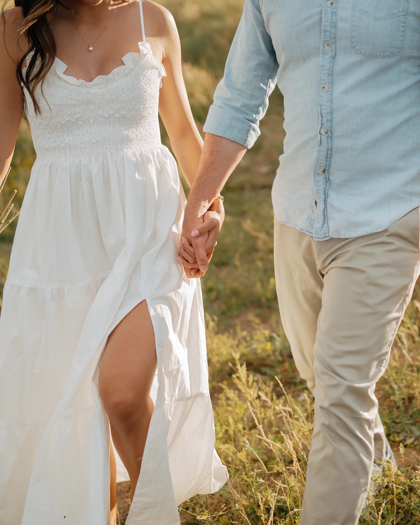 A woman in a white dress holding hands with a man in light-colored pants and a button-up shirt, walking outdoors on a grassy field.