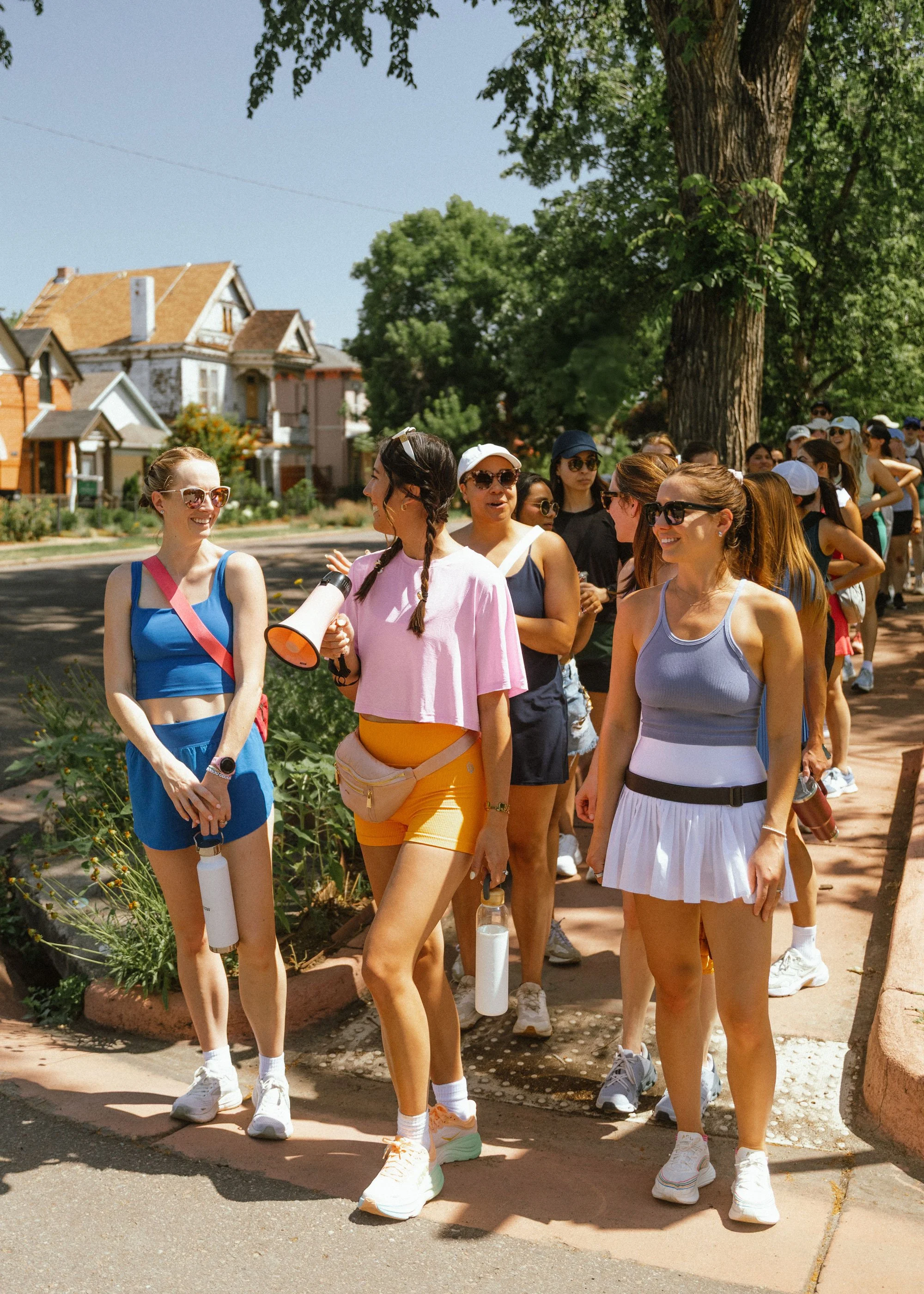 Group of women standing in a line on a sidewalk outdoors, some wearing athletic clothes and sunglasses, with trees and houses in the background.