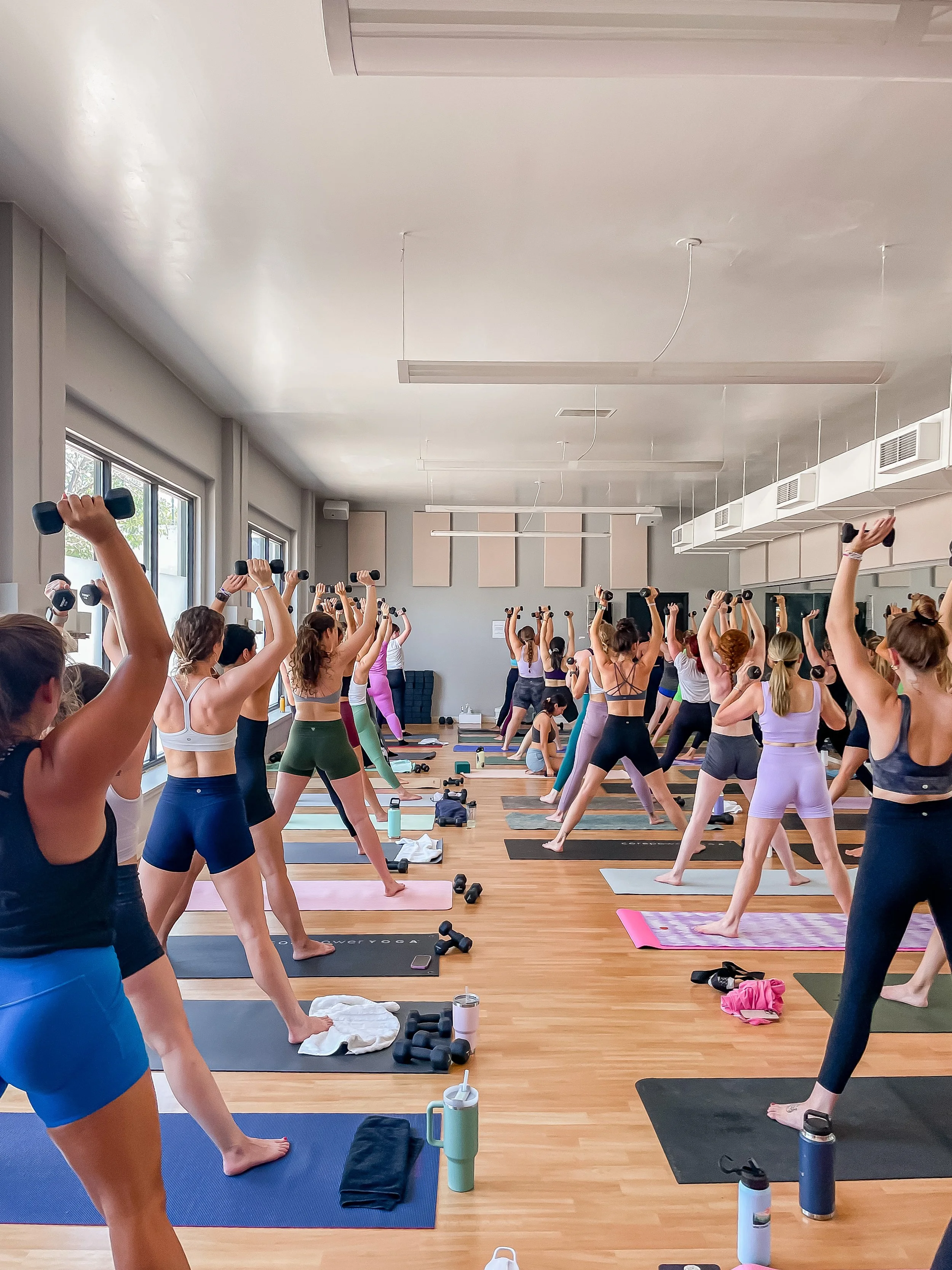 A group of women participating in a yoga class in a bright studio, raising dumbbells overhead, with yoga mats and water bottles on the floor.