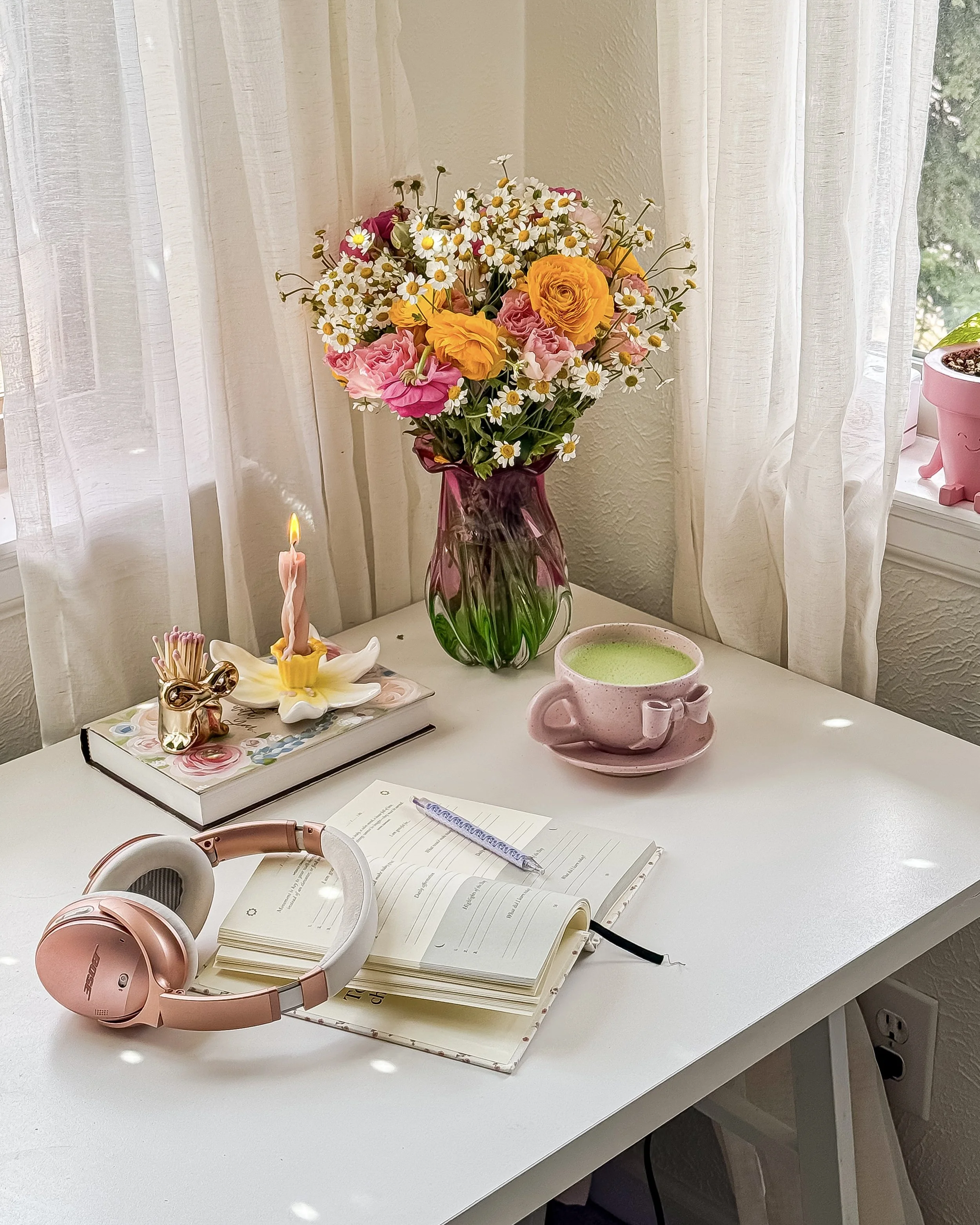 A white desk with a bouquet of mixed flowers in a pink vase, a lit pink candle, a pink mug with a green beverage, a notebook with a pen, a pair of pink over-ear headphones, and a small ceramic figurine.