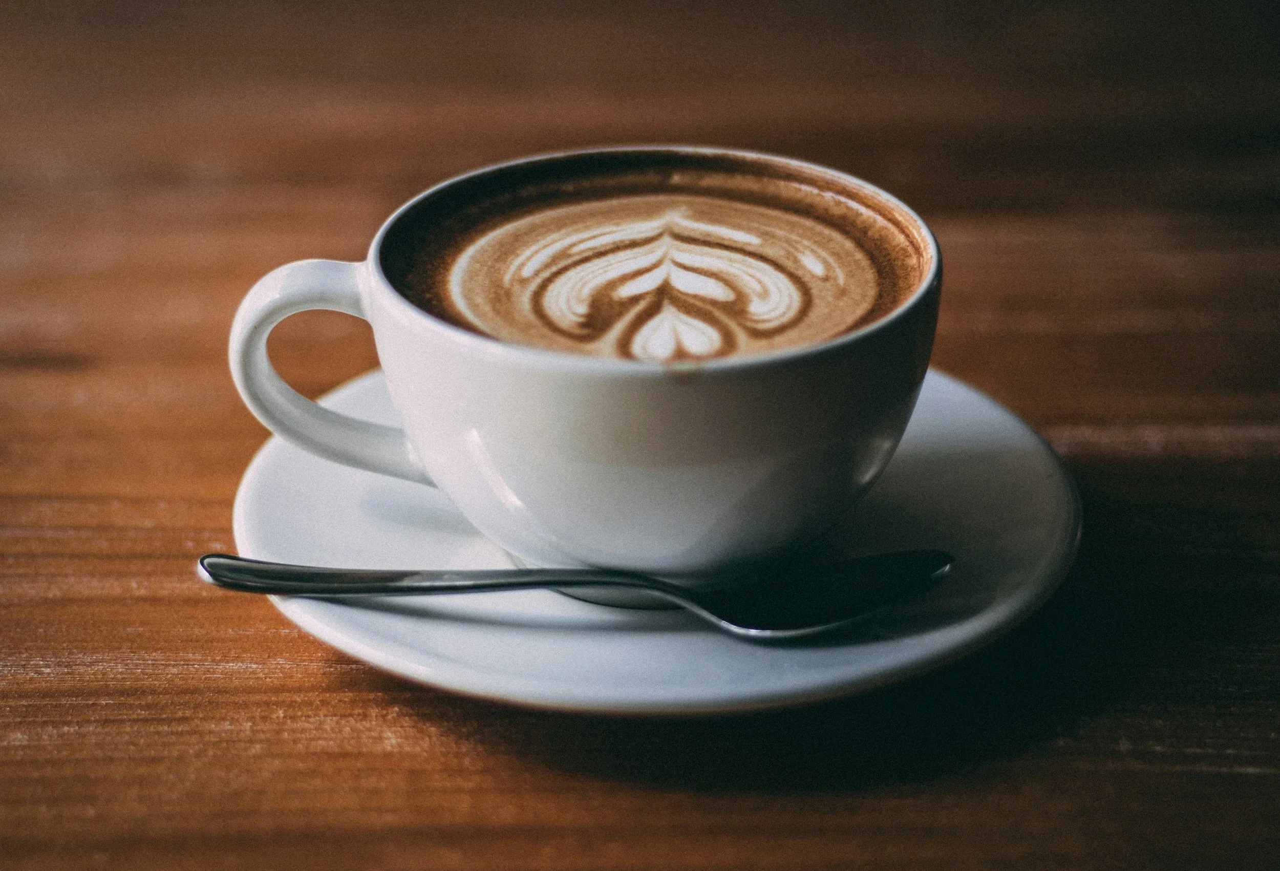White cup of coffee with latte art on top, placed on a white saucer with a spoon, on a wooden table.