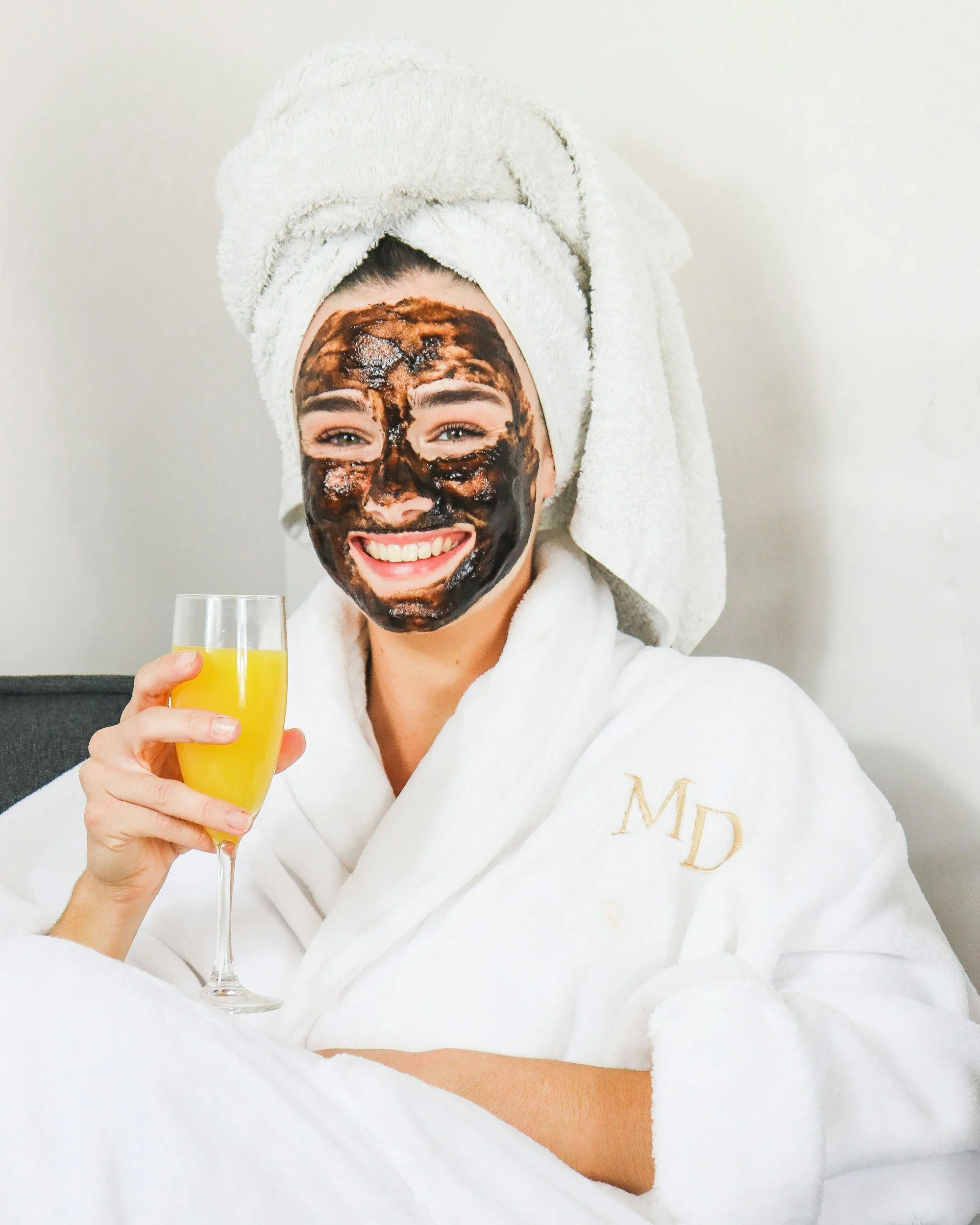 A woman with a towel wrapped around her head and a bathrobe with the initials 'MD' embroidered on it, smiling and holding a glass of orange juice. She has a charcoal facial mask on her face.