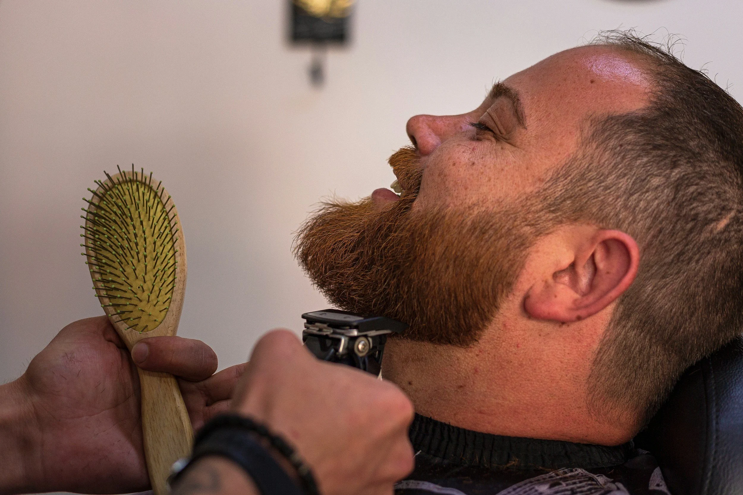 A man with a full beard is getting shaved with an electric razor, leaning back against a chair. A person is holding a wooden beard brush close to his face.