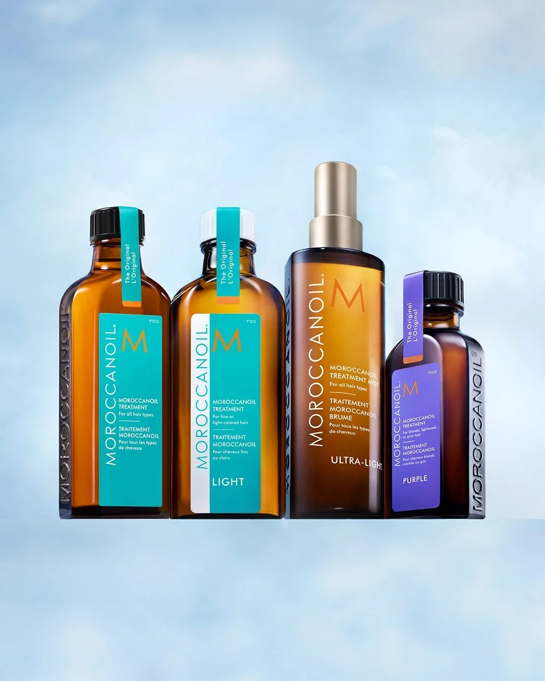 Four bottles of Moroccanoil hair treatment products in various sizes and colors, set against a light blue background.