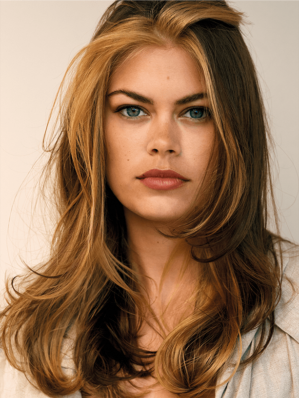 A woman with long, wavy hair with blonde and brown highlights, wearing a beige top, and looking directly at the camera.