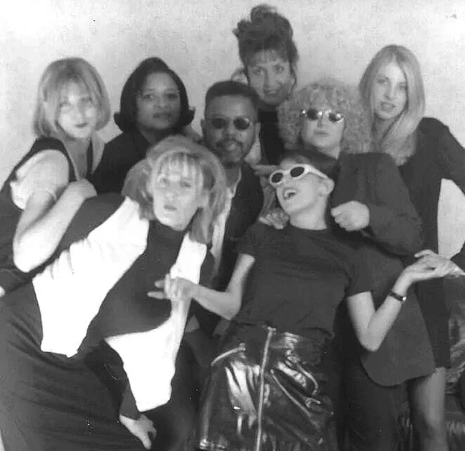 Group of eight people posing together, with some wearing sunglasses and trendy clothing, in black and white.