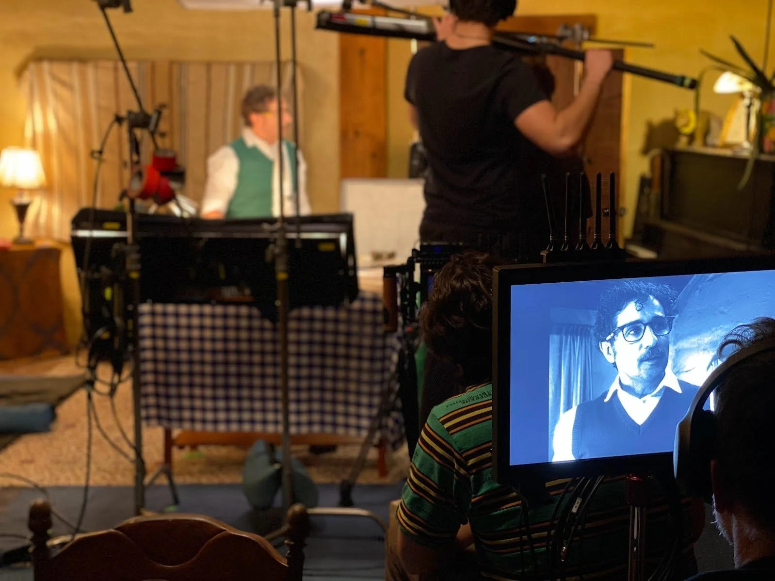 Behind-the-scenes of a film or video production with a monitor displaying a black-and-white image of a man with glasses, and crew members setting up equipment in a cozy, rustic room.