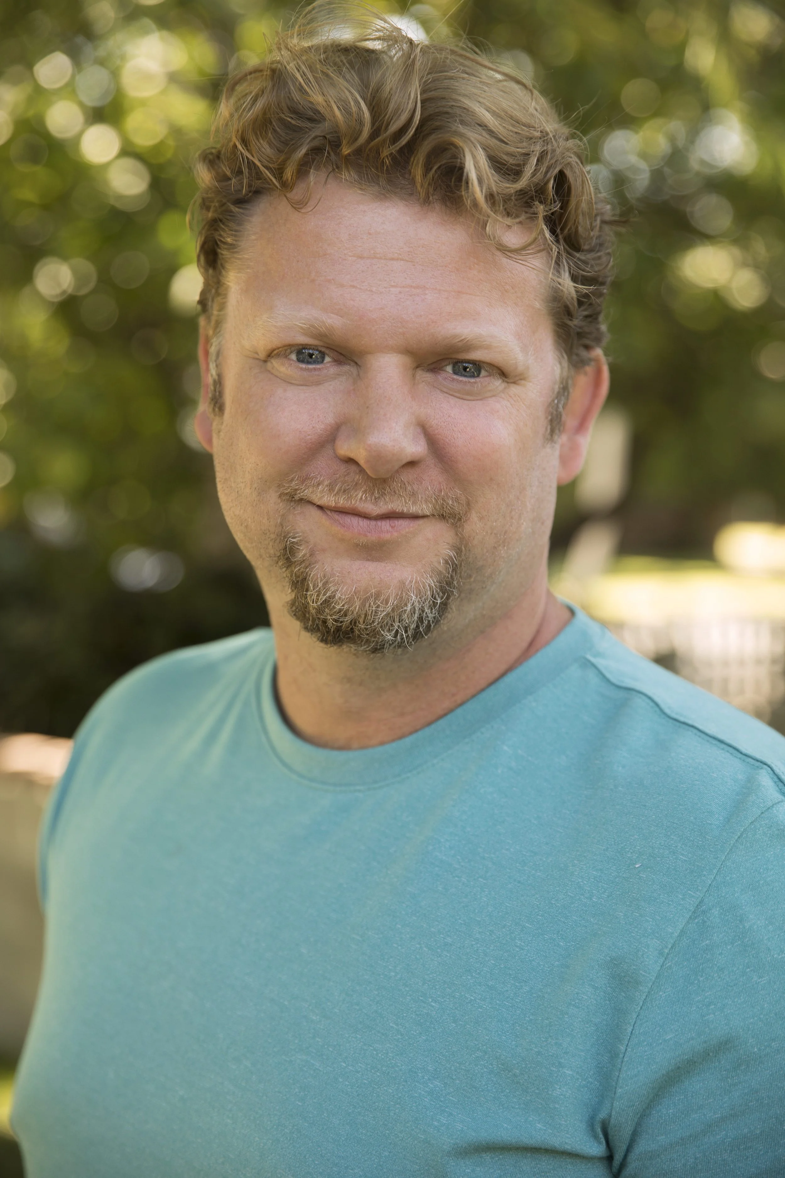 A man with curly light brown hair, blue eyes, and a beard, wearing a turquoise t-shirt, smiling outdoors with green trees and sunlight in the background.