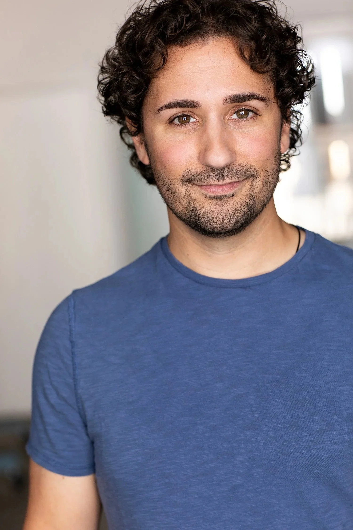 Headshot of a smiling man with curly brown hair, brown eyes, light skin, wearing a blue t-shirt, against a blurred light background.