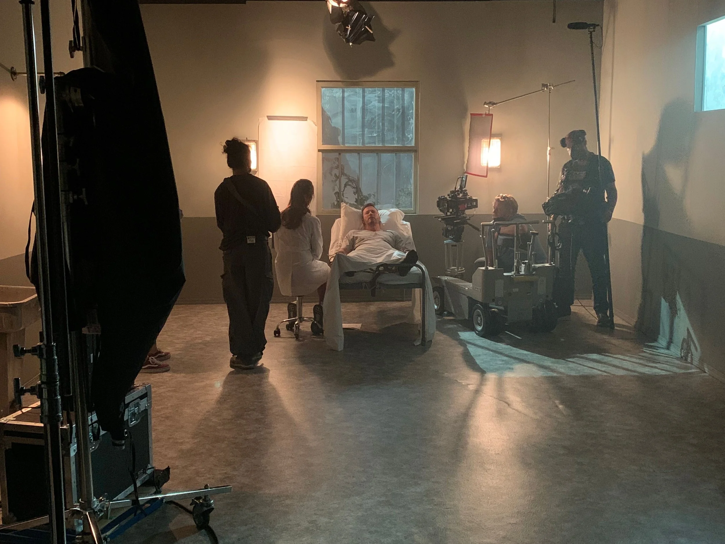 A film set with a hospital scene, featuring a man lying in a hospital bed, surrounded by crew members, camera operators, and lighting equipment in a dimly lit room with windows.