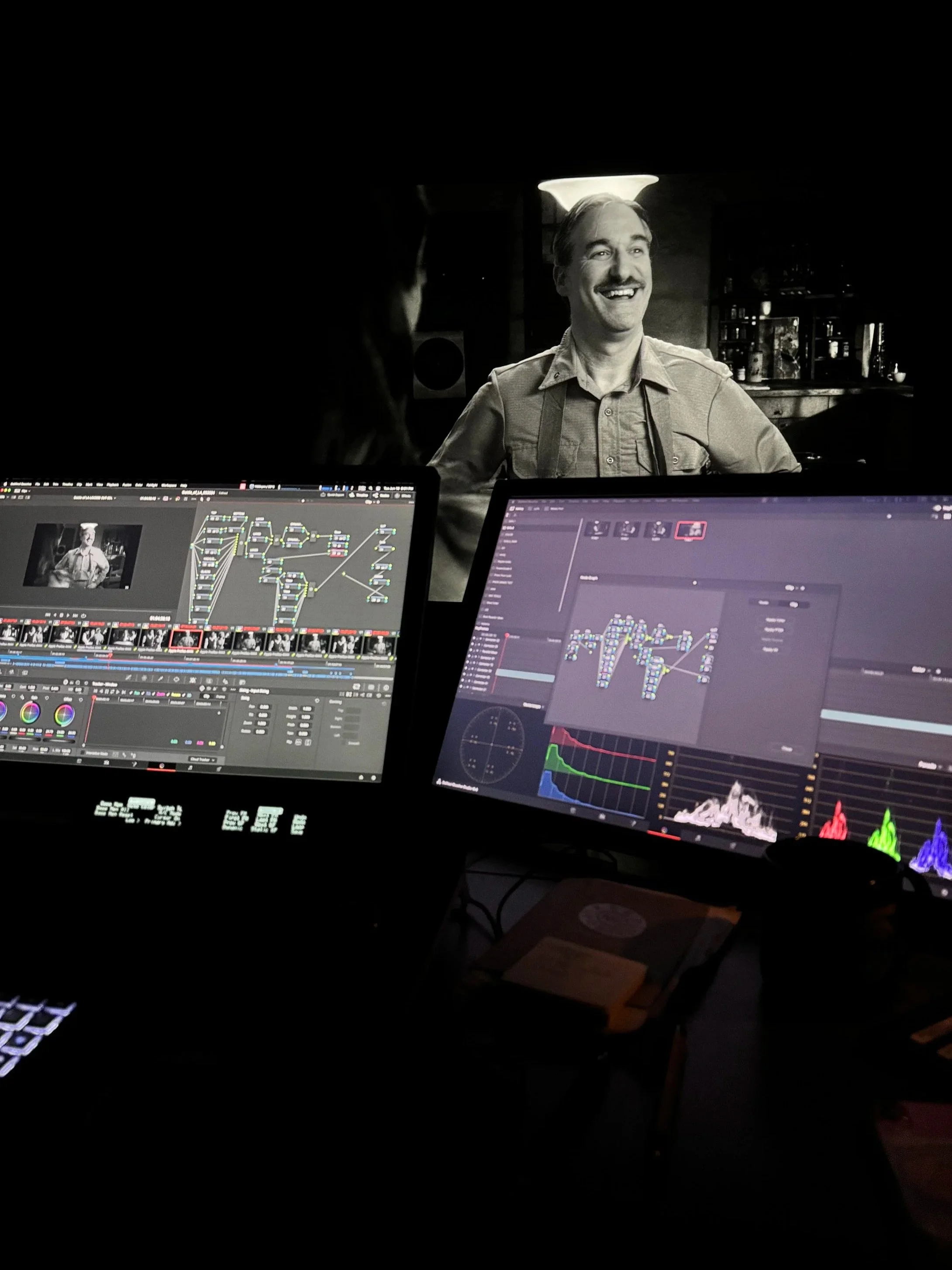 Black and white photo of a smiling man with a mustache sitting at a bar or restaurant, with two widescreen computer monitors displaying video editing software in the foreground.