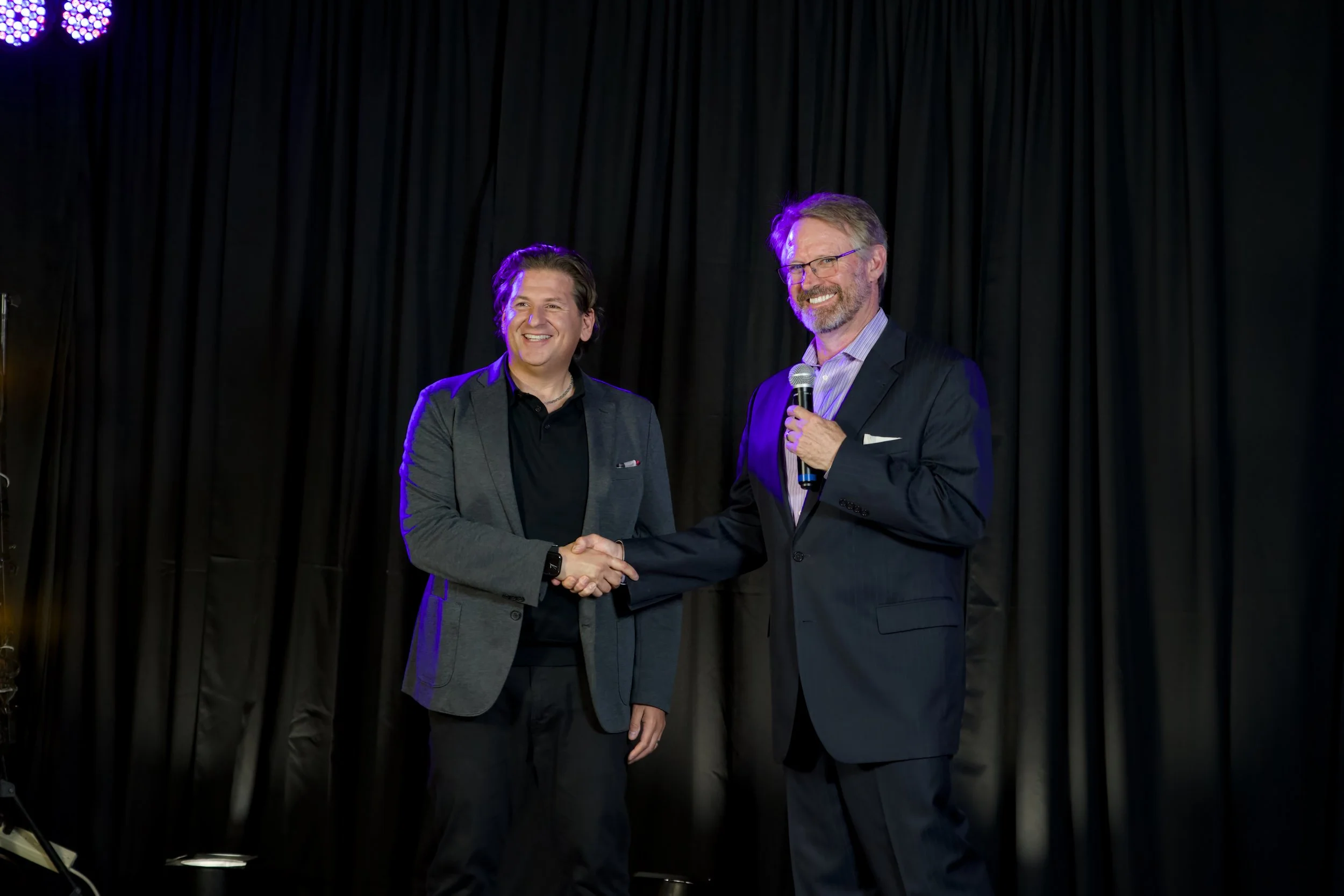 Two men on stage shaking hands, one holding a microphone, with a black curtain backdrop.