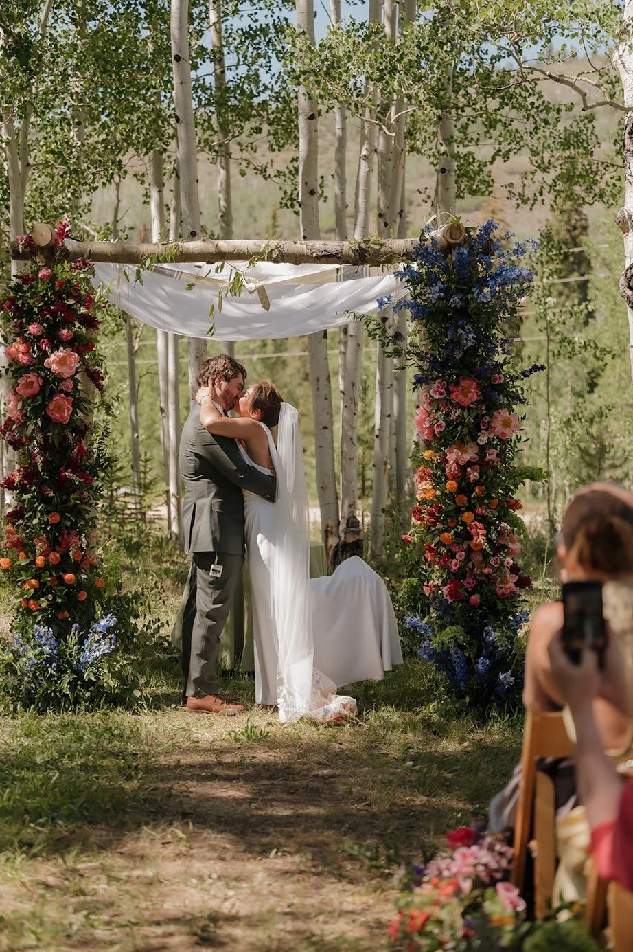A bride and groom kiss under a luxury wedding arch decorated with colorful flowers in a forest setting.