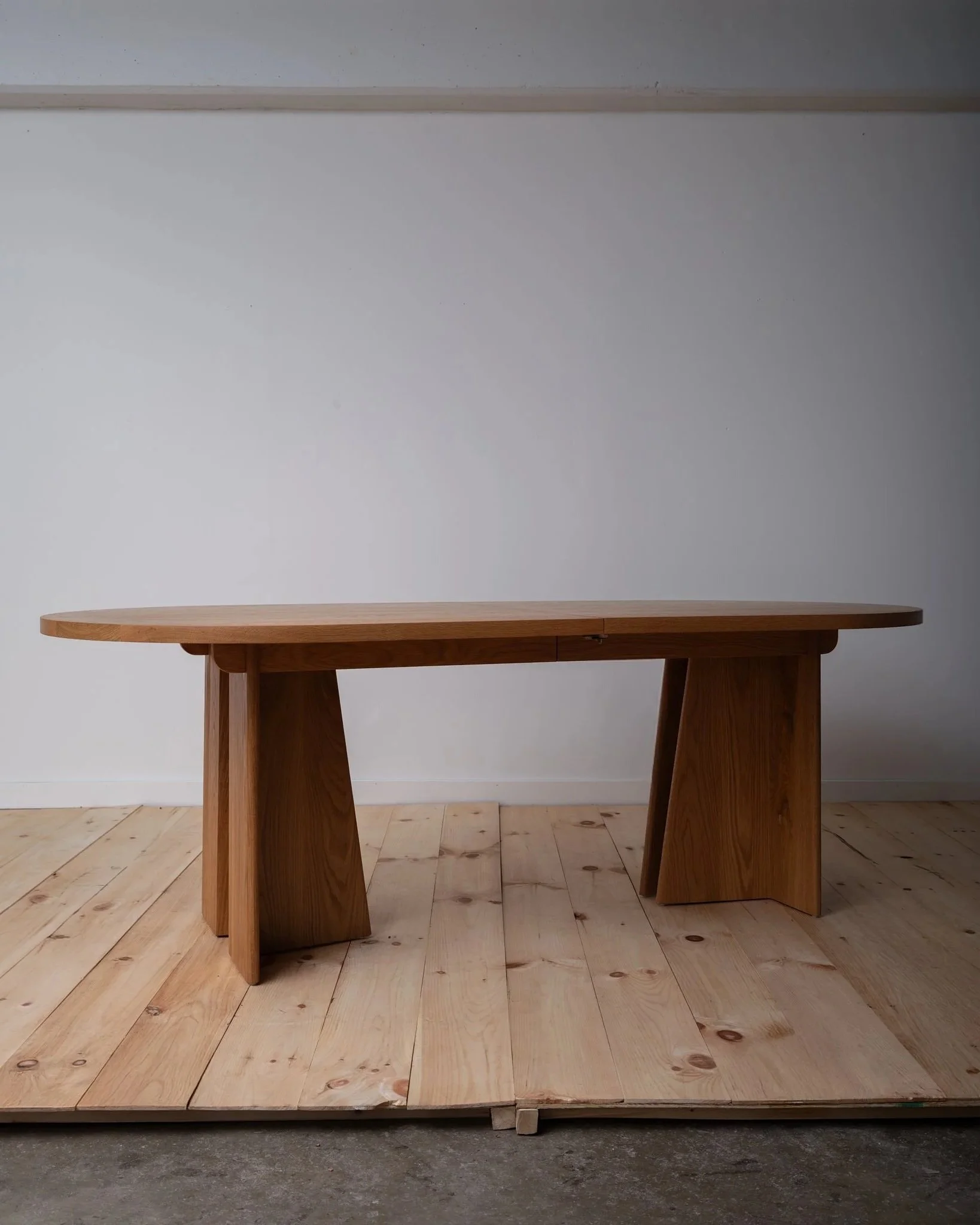 A wooden table with a curved rectangular top and wide, angled legs, placed on a wooden floor against a white wall.