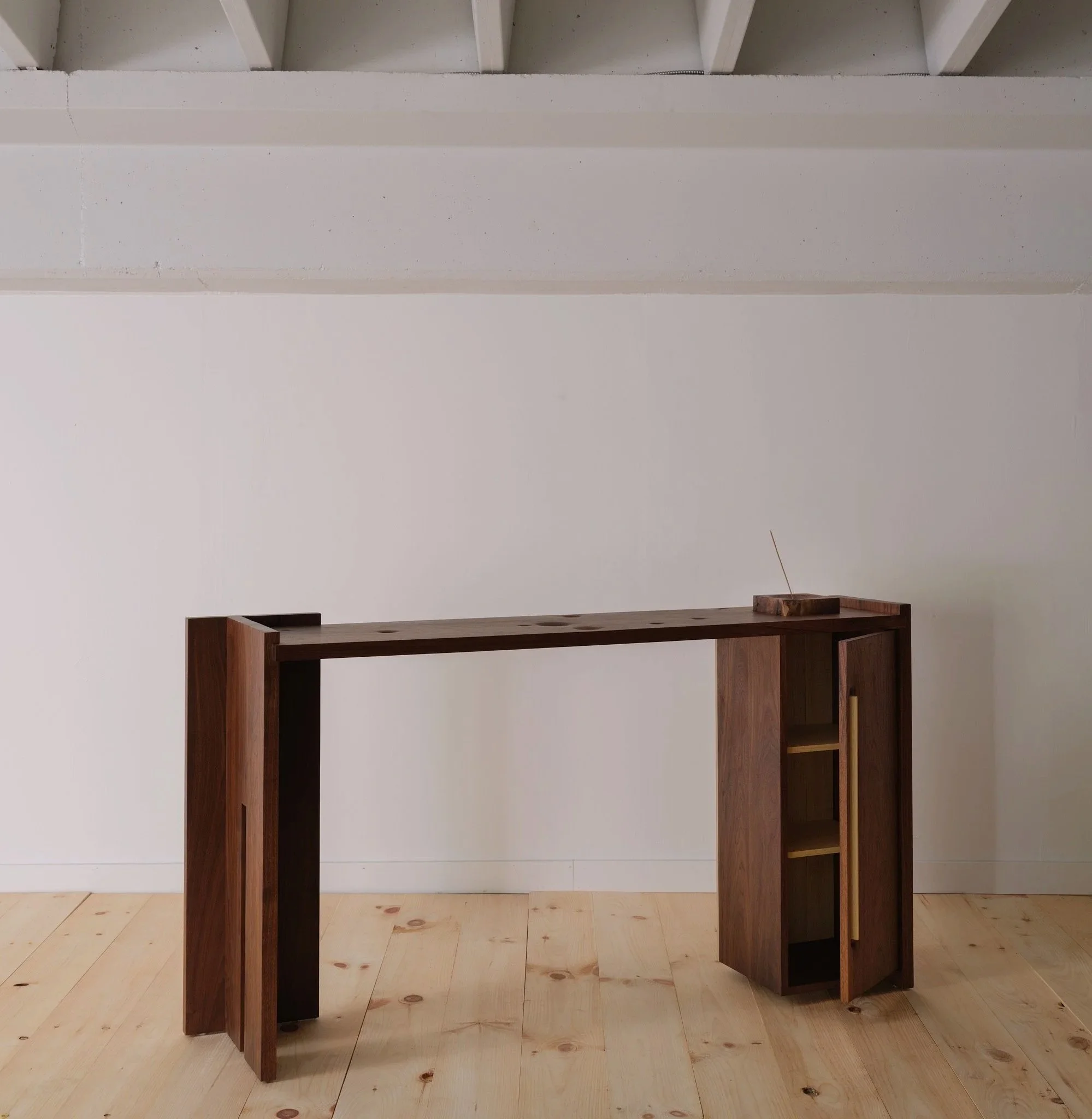 A wooden desk with open shelving on the right side, situated on a light wooden floor against a plain white wall.