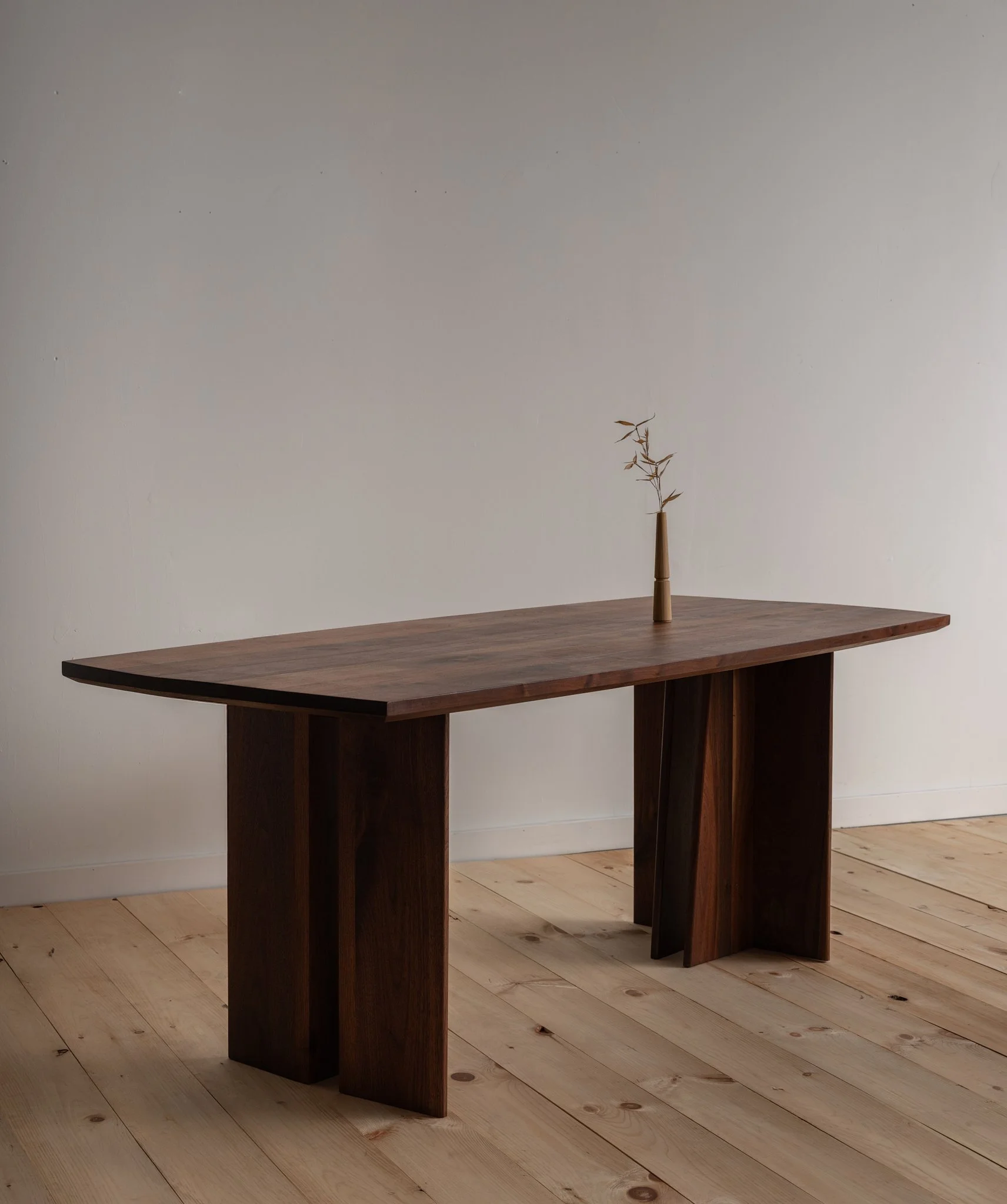 A minimalist wooden table with a single small, bare, dry branch in a narrow vase on top, set against a blank, off-white wall and a natural wood floor.