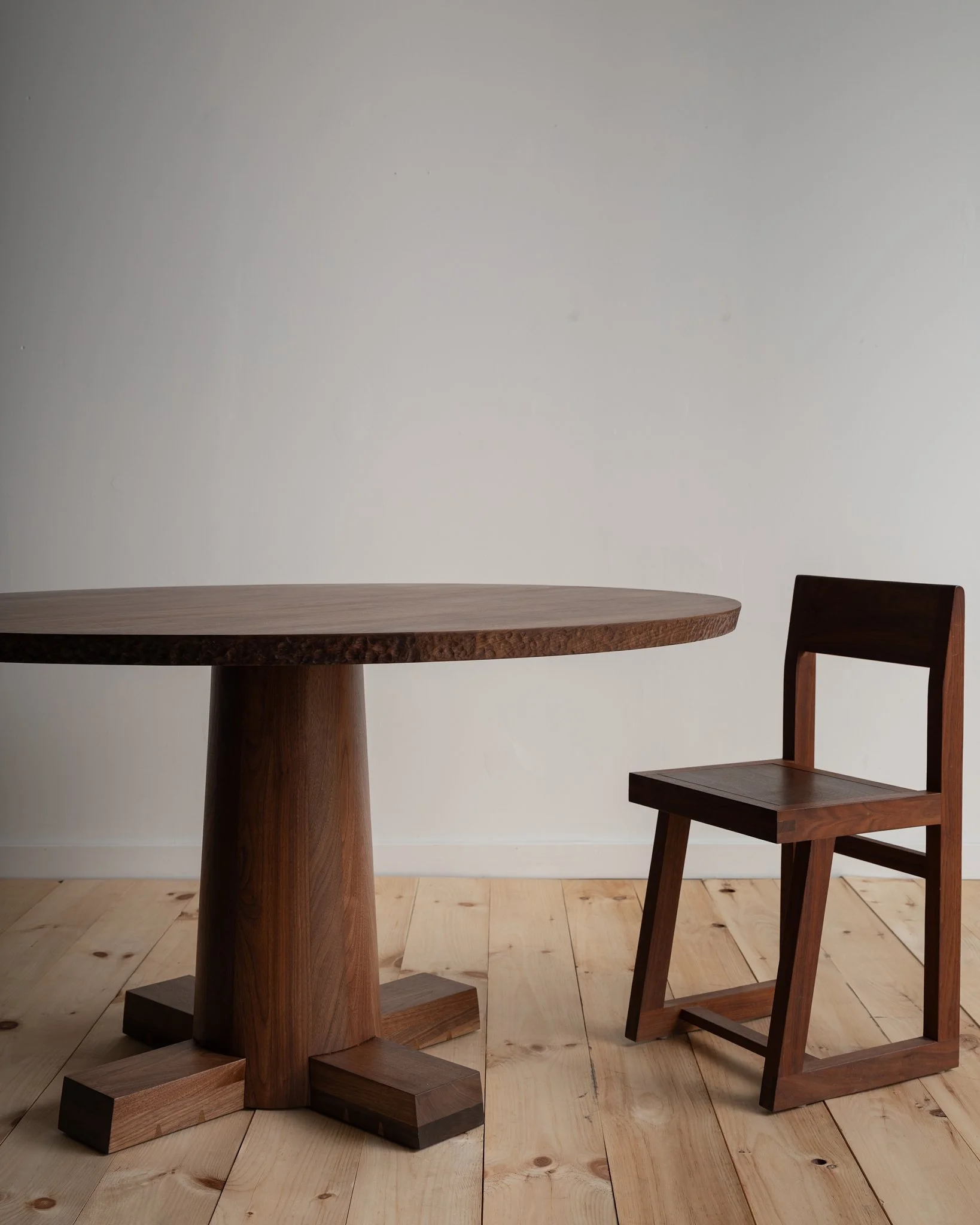 A wooden round table and a matching wooden chair in a minimalistic room with light wooden flooring and a plain white wall.