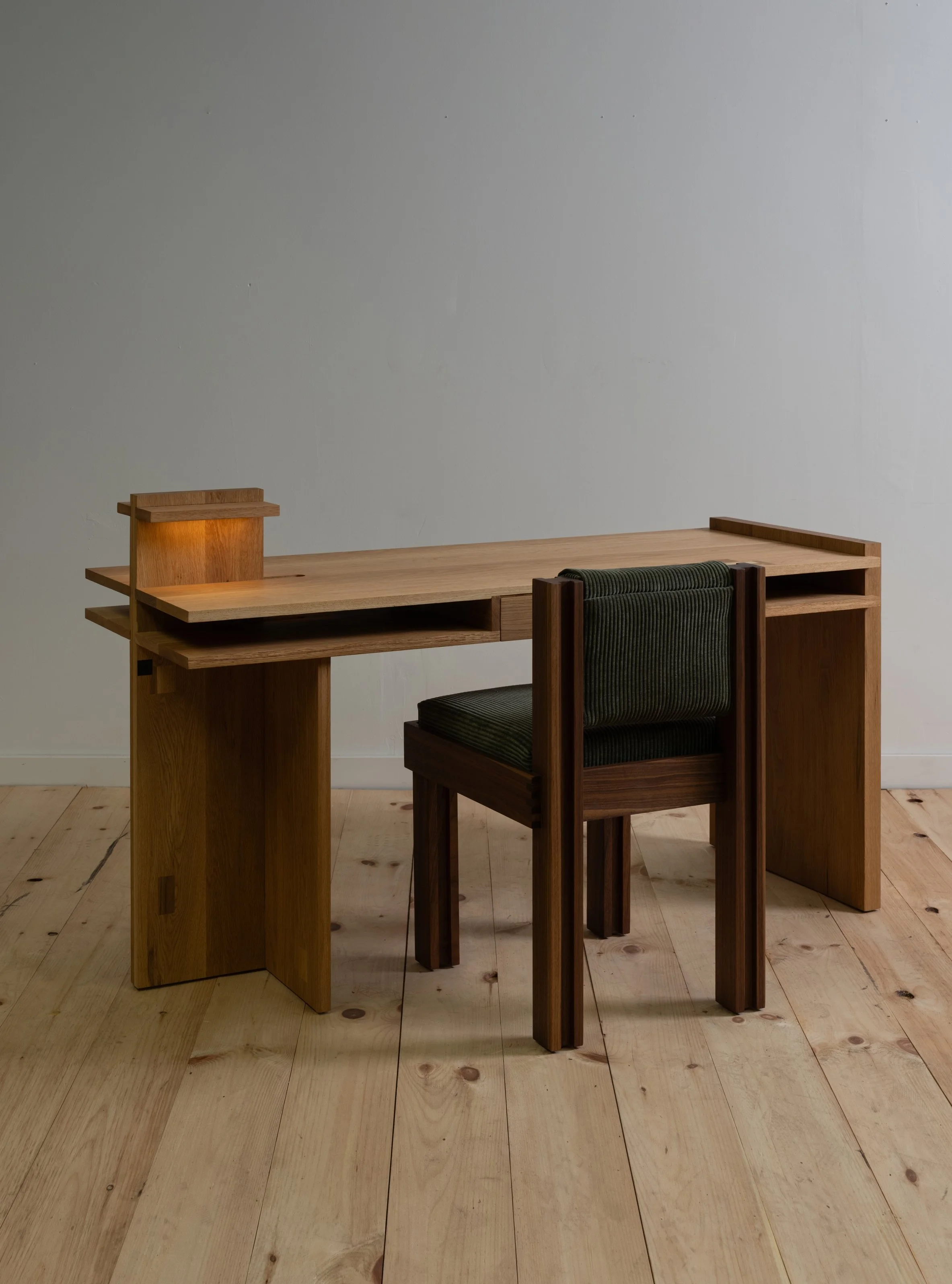 A wooden desk with a small raised platform and a matching wooden chair in a room with light-colored wooden floors and a plain light gray wall.