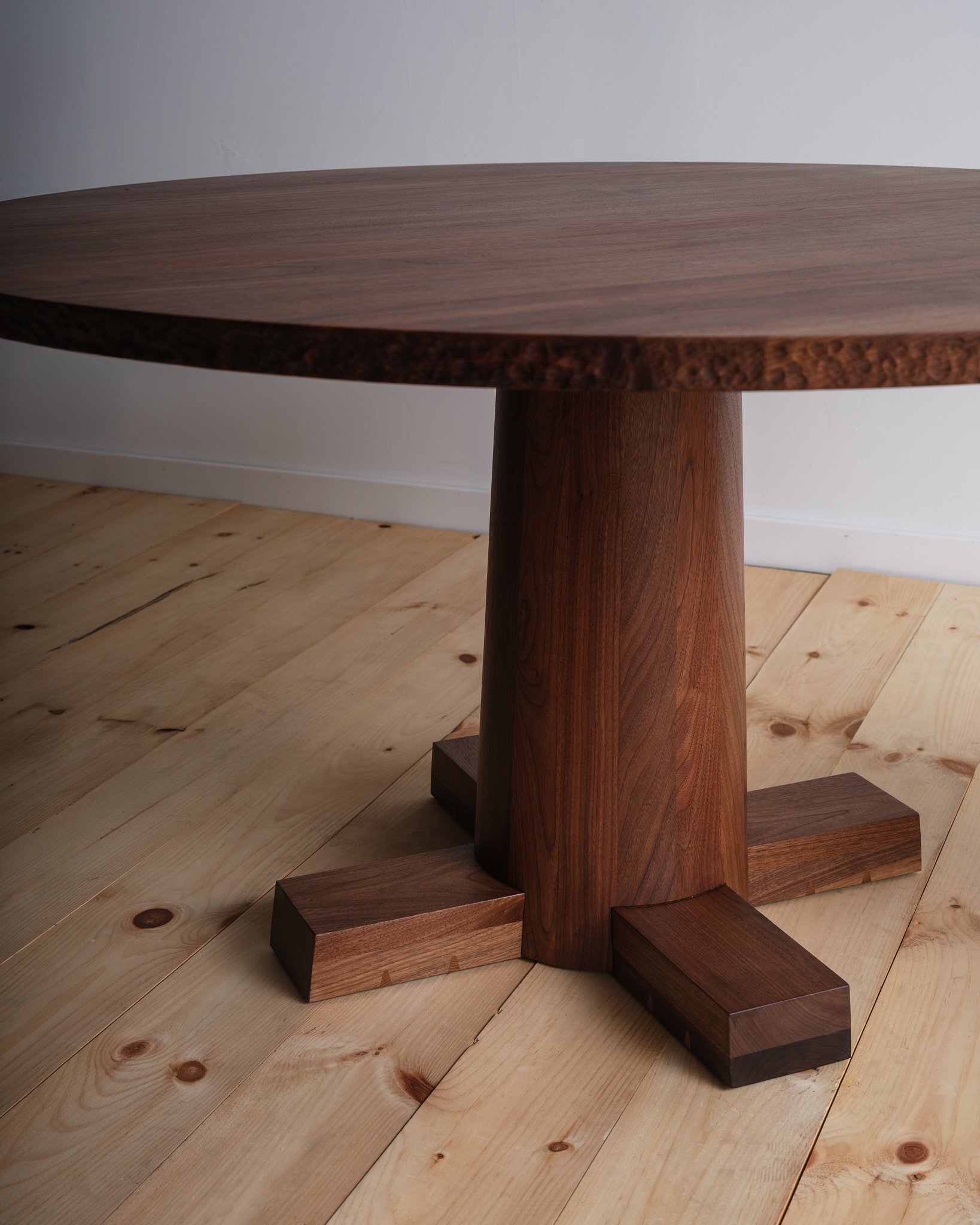 Close-up of a wooden round table with a central pedestal base and four supports, on a light wooden floor against a white wall.