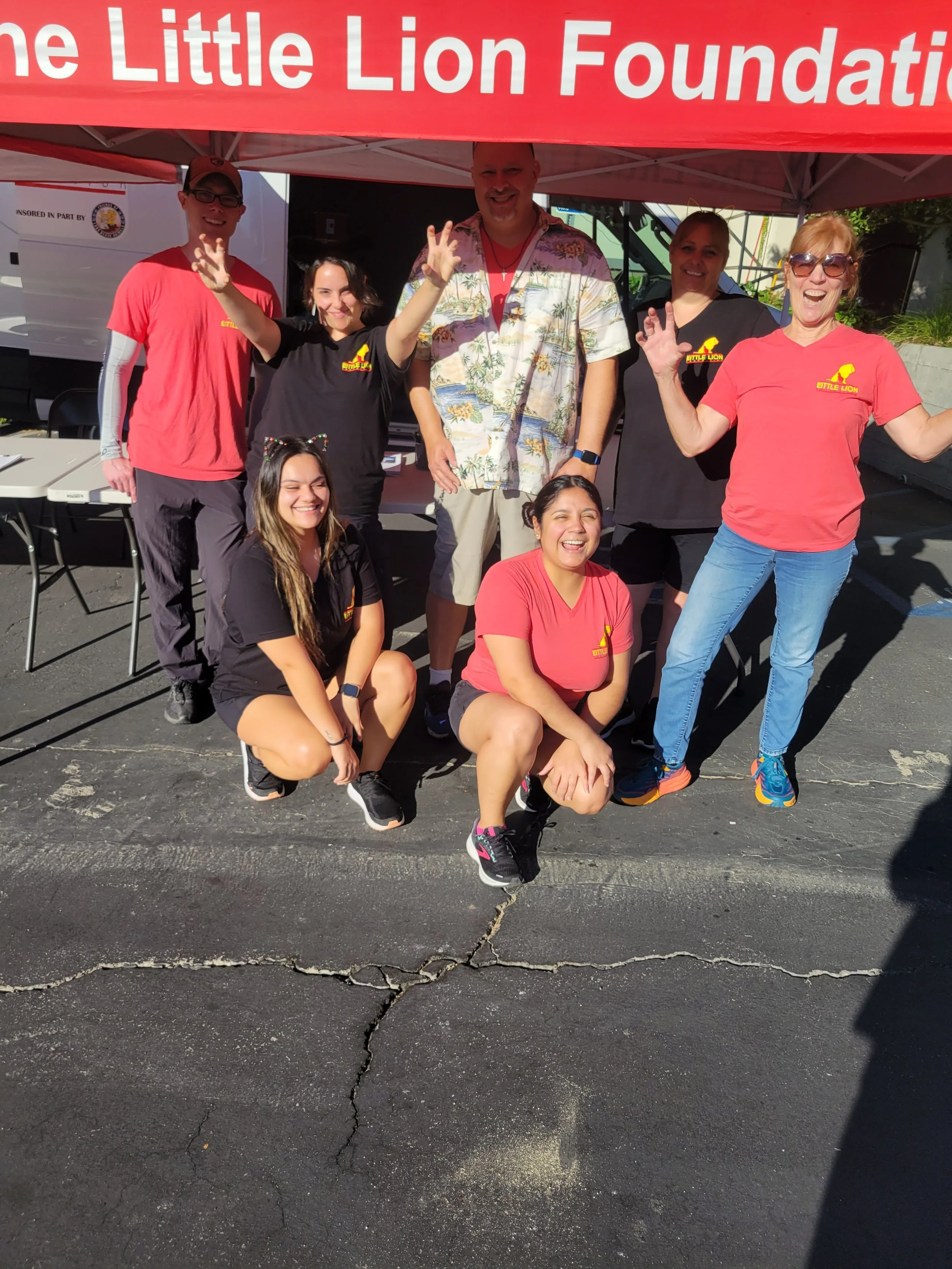 Group of people smiling and posing in front of a tent with a sign that reads 'The Little Lion Foundation'.