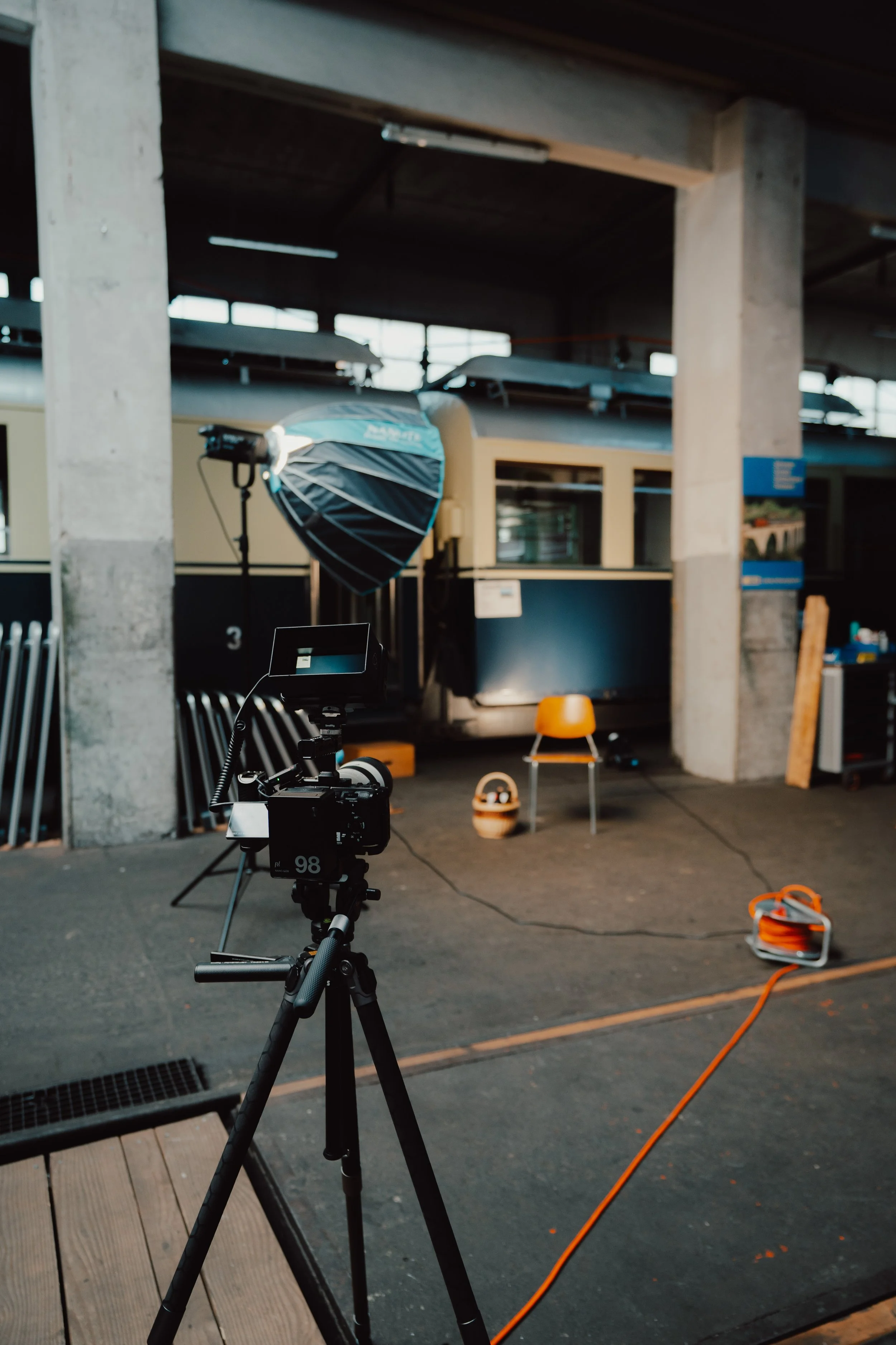 Innenaufnahme eines Fotostudios mit Kamera auf Stativ, Beleuchtung, Stuhl, Kabel, und Hintergründen für Fotoshootings.