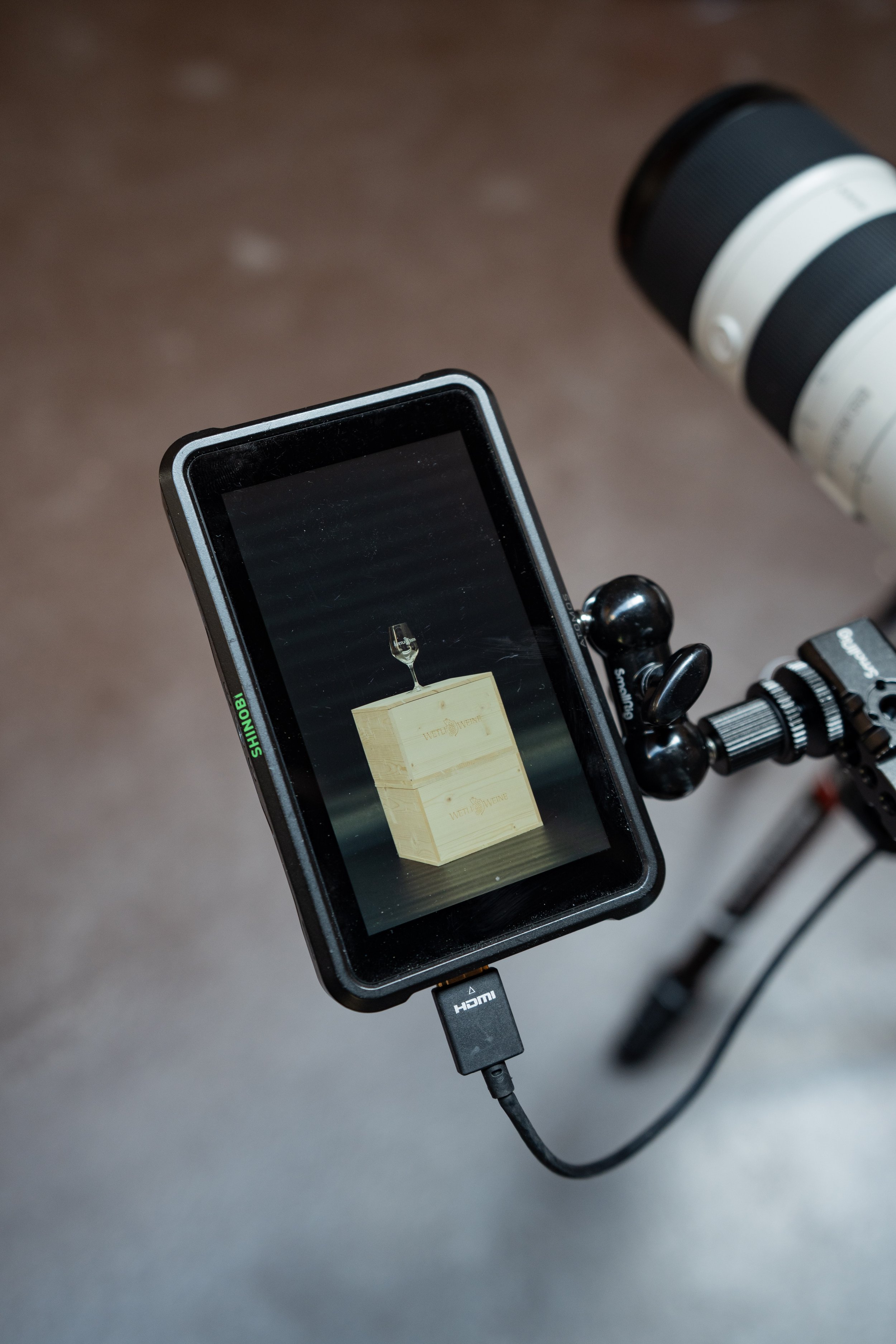 Ein Smartphone auf einem Stativ zeigt ein Bild von einem Holzkasten mit einem Glaselement oben darauf. Im Hintergrund befindet sich ein Kamerobjektiv.