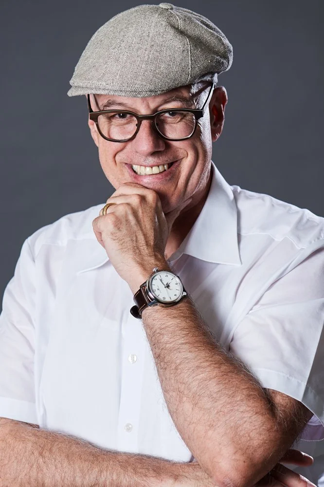 Ein Mann trägt eine weiße Hemd und eine graue Mütze. Er trägt eine Brille, hat einen goldenen Ehering an der Hand und trägt eine Armbanduhr. Er lächelt und hat die Hand am Kinn.