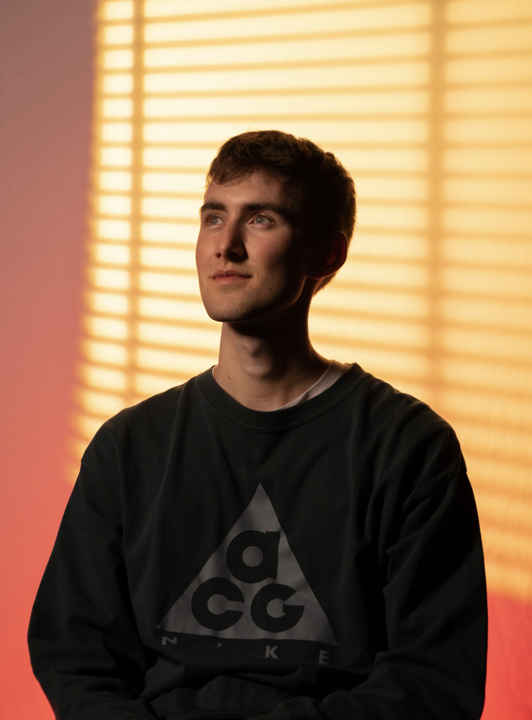 Ein junger Mann mit braunen Haaren und blauen Augen sitzt vor einer warm beleuchteten Jalousie, trägt ein schwarzes Sweatshirt mit einem geometrischen Logo.