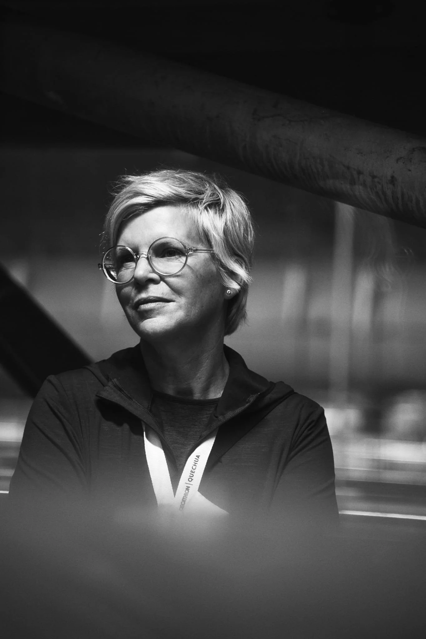 Schwarz-weiß-Foto einer Frau mit kurzen, blonden Haaren und Brille, die nach links schaut, getragen eine dunkle Jacke, im Hintergrund eine Industrielandschaft.