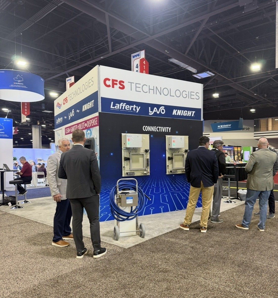 A trade show booth for CFS Technologies with a blue digital circuit design backdrop, promoting connectivity products and brands Lafferty, YAVO, and Knight. Several people are engaged in conversation around the booth.