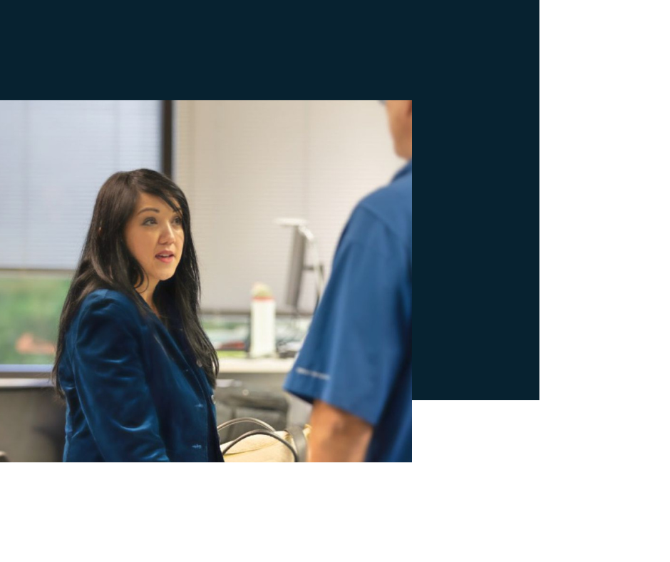A woman with dark brown hair, wearing a blue blazer, talking to a man in a blue uniform in a hospital or medical setting.