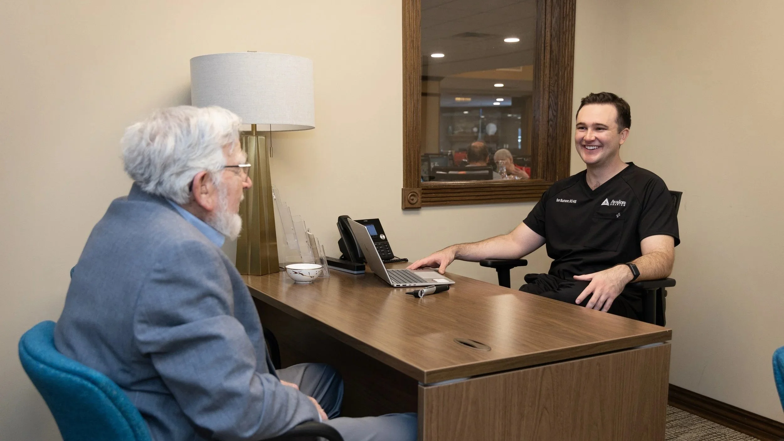 A young male healthcare professional sitting at a desk with a laptop, engaging in conversation with an older man in a professional setting.