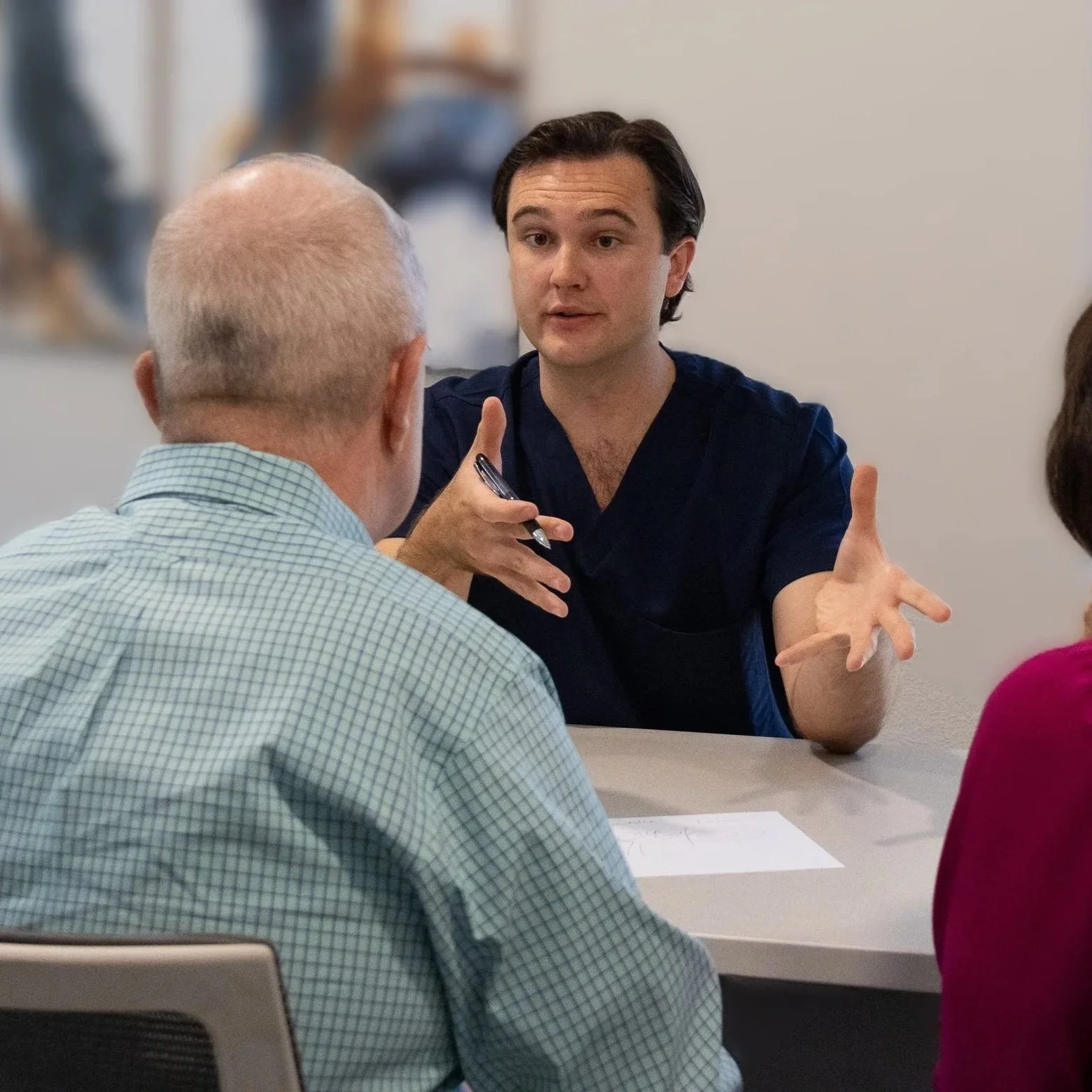 A young man wearing navy scrubs engaged in a conversation with an older man and woman, gesturing with his hands as he speaks.