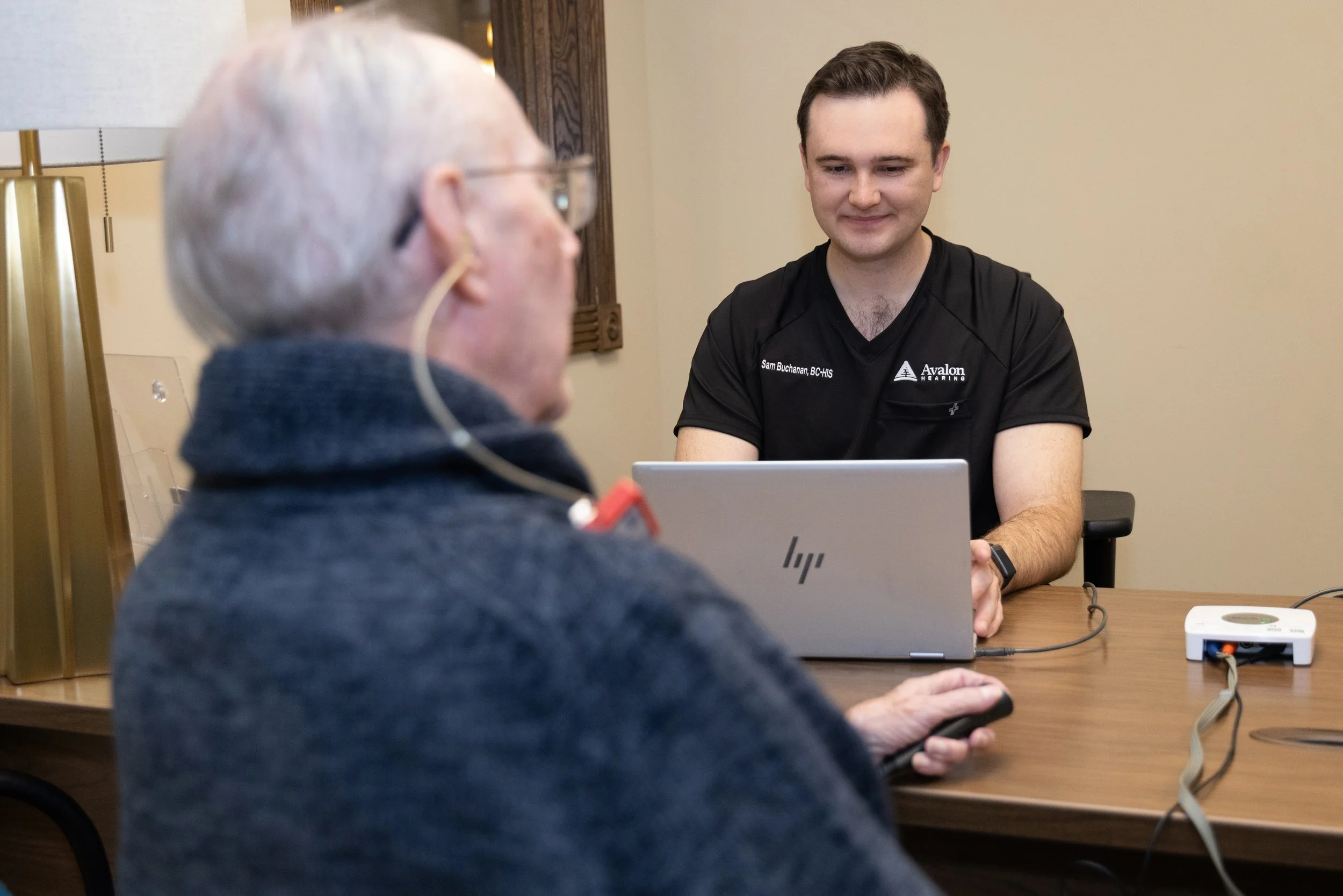 A young man wearing a black shirt is sitting at a table with a laptop, talking to an older person with a hearing aid, who is holding a remote control device.