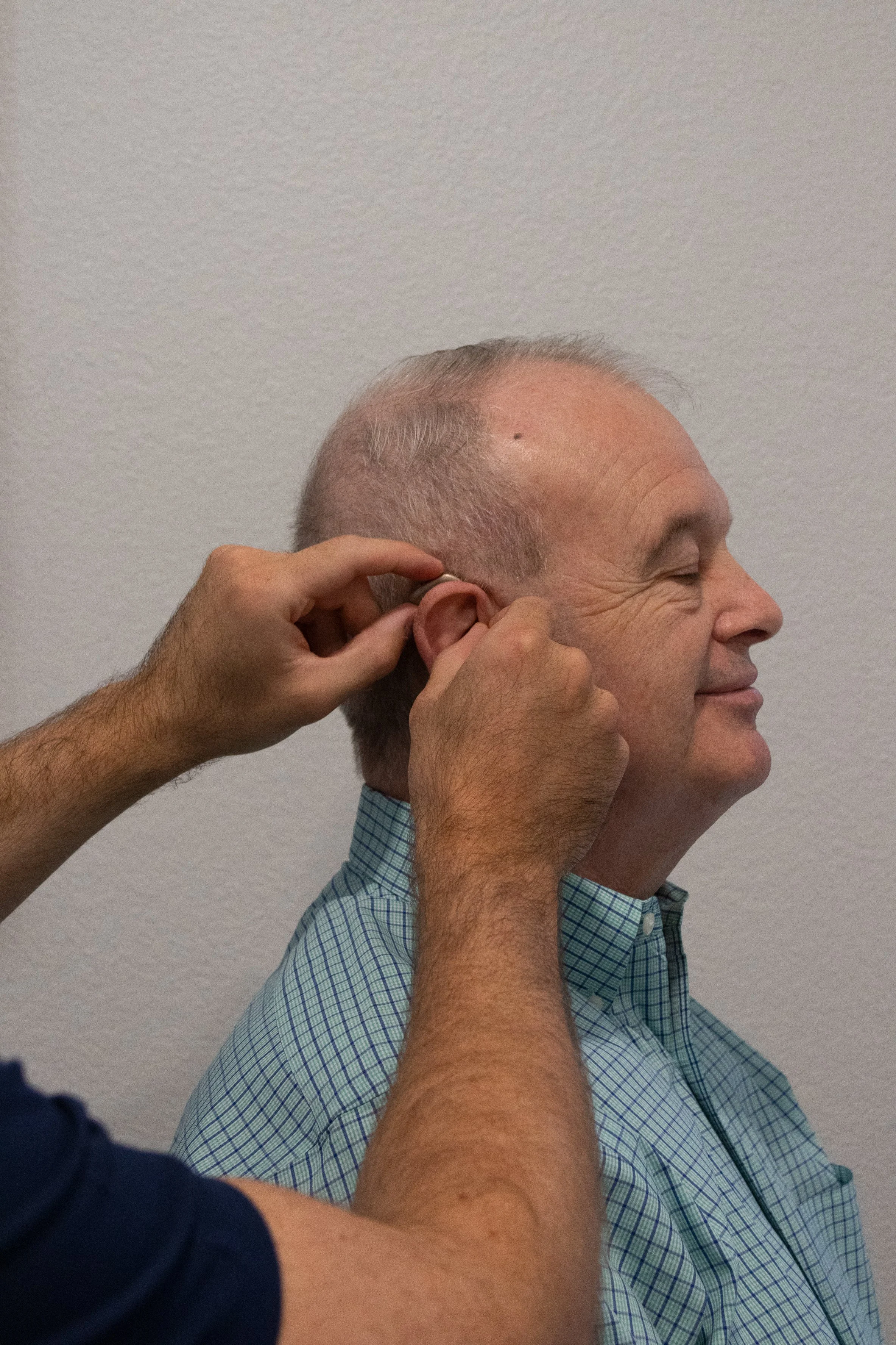 A man is sitting with his eyes closed as another person inserts a hearing aid into his ear. The man is wearing a blue checkered shirt and is smiling gently.