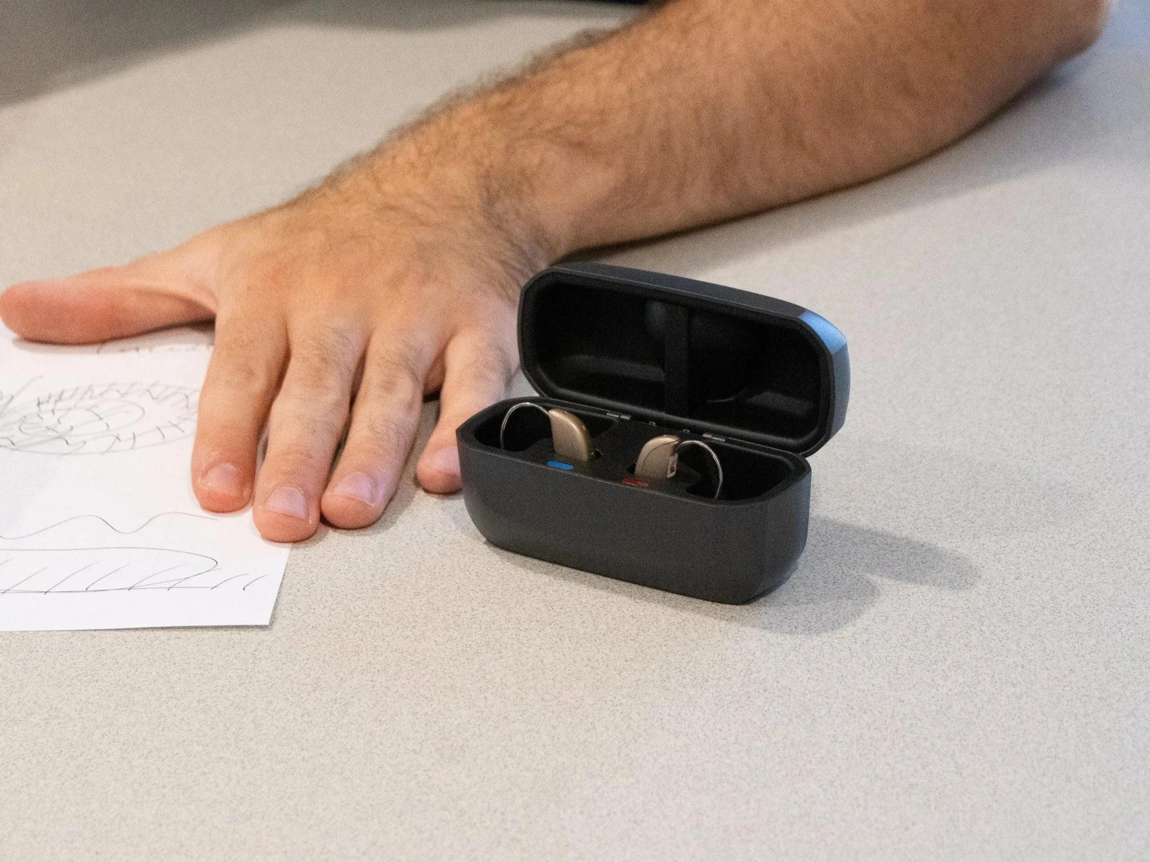 A person's arm and hand resting on a gray table next to a black case with open lid, containing a pair of hearing aids inside.