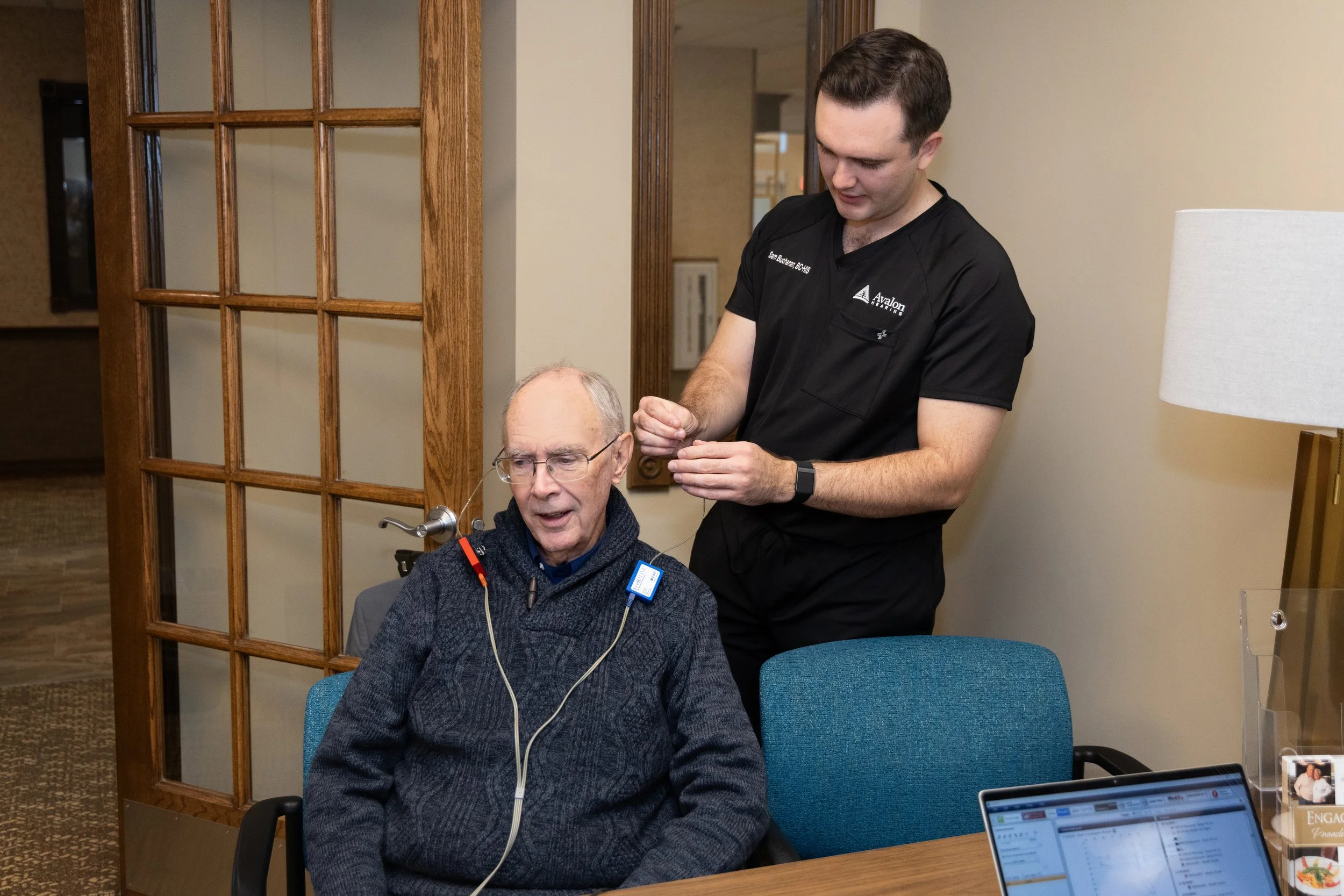 A young man in black medical scrubs assisting an elderly man with a hearing aid, sitting at a table in a room with wood details.