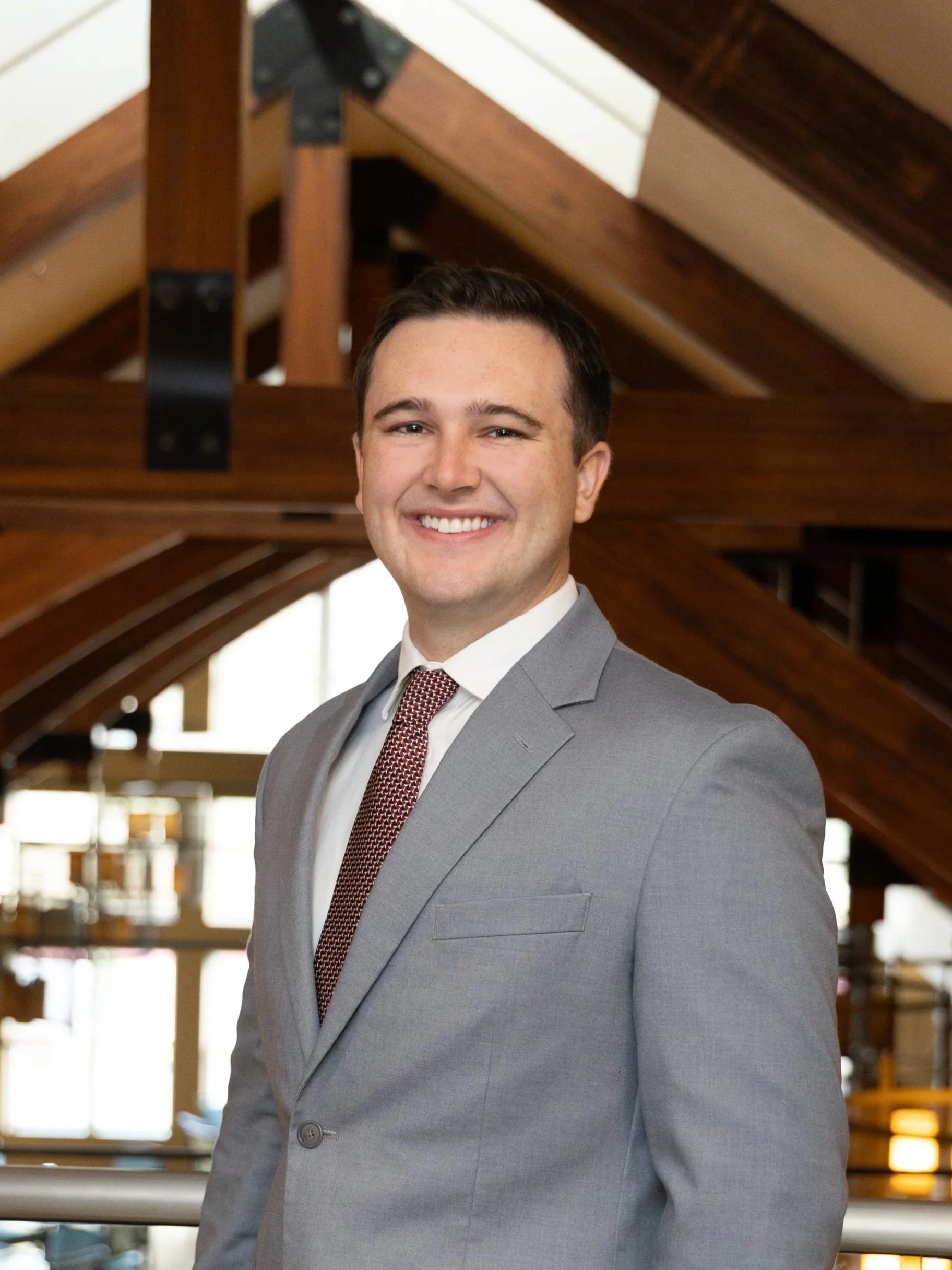A young man in a gray business suit, white shirt, and patterned red tie standing inside a building with wooden beams in the background, smiling at the camera.
