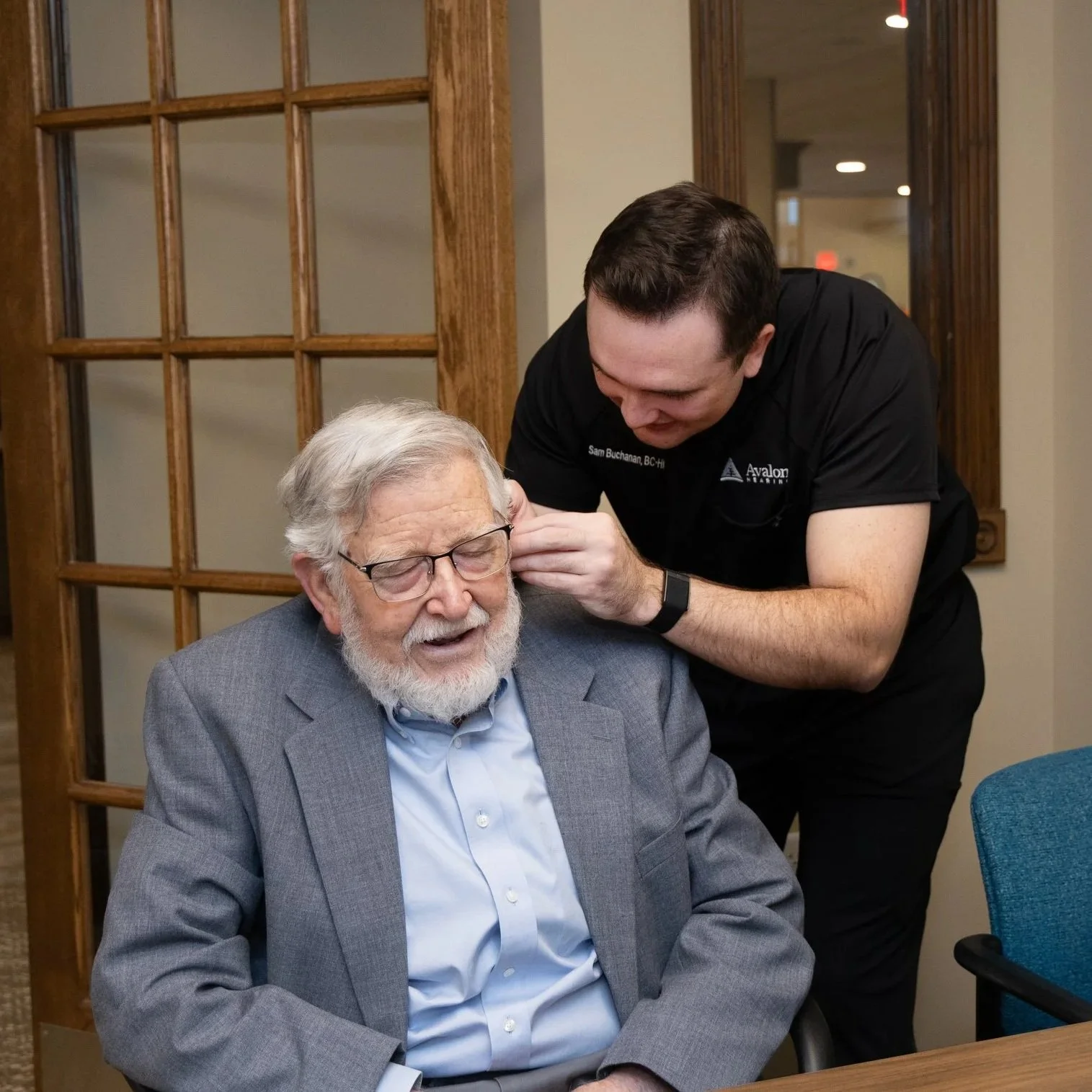 An elderly man with glasses and a gray suit is smiling with his eyes closed as a young man helps him put on hearing aids behind his ears in a room with wooden framed glass doors.