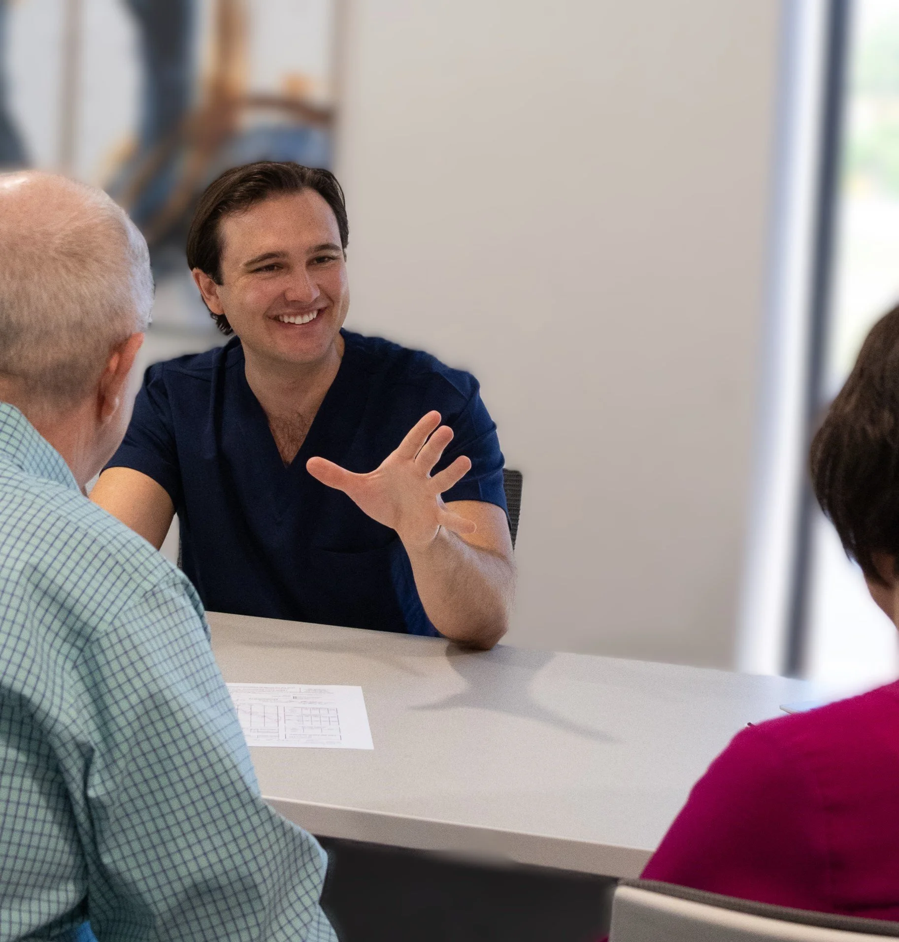 A young man in a navy blue medical scrubs smiling and gesturing with his hand while talking to two older adults, a man with a bald head and a woman with short dark hair, sitting at a table in a room with natural light.