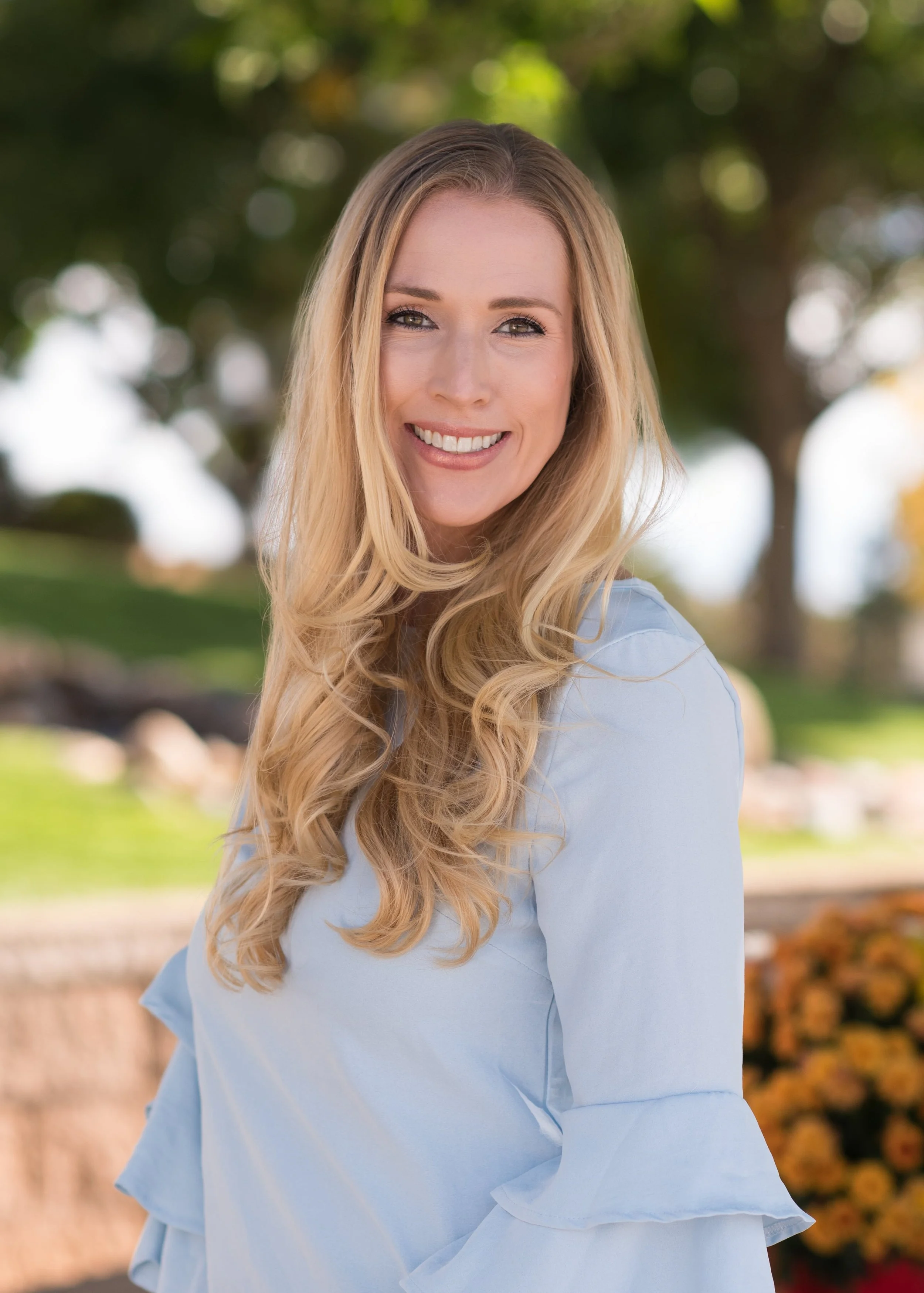 A smiling woman with long blonde hair wearing a light blue dress outdoors with trees and flowers in the background.